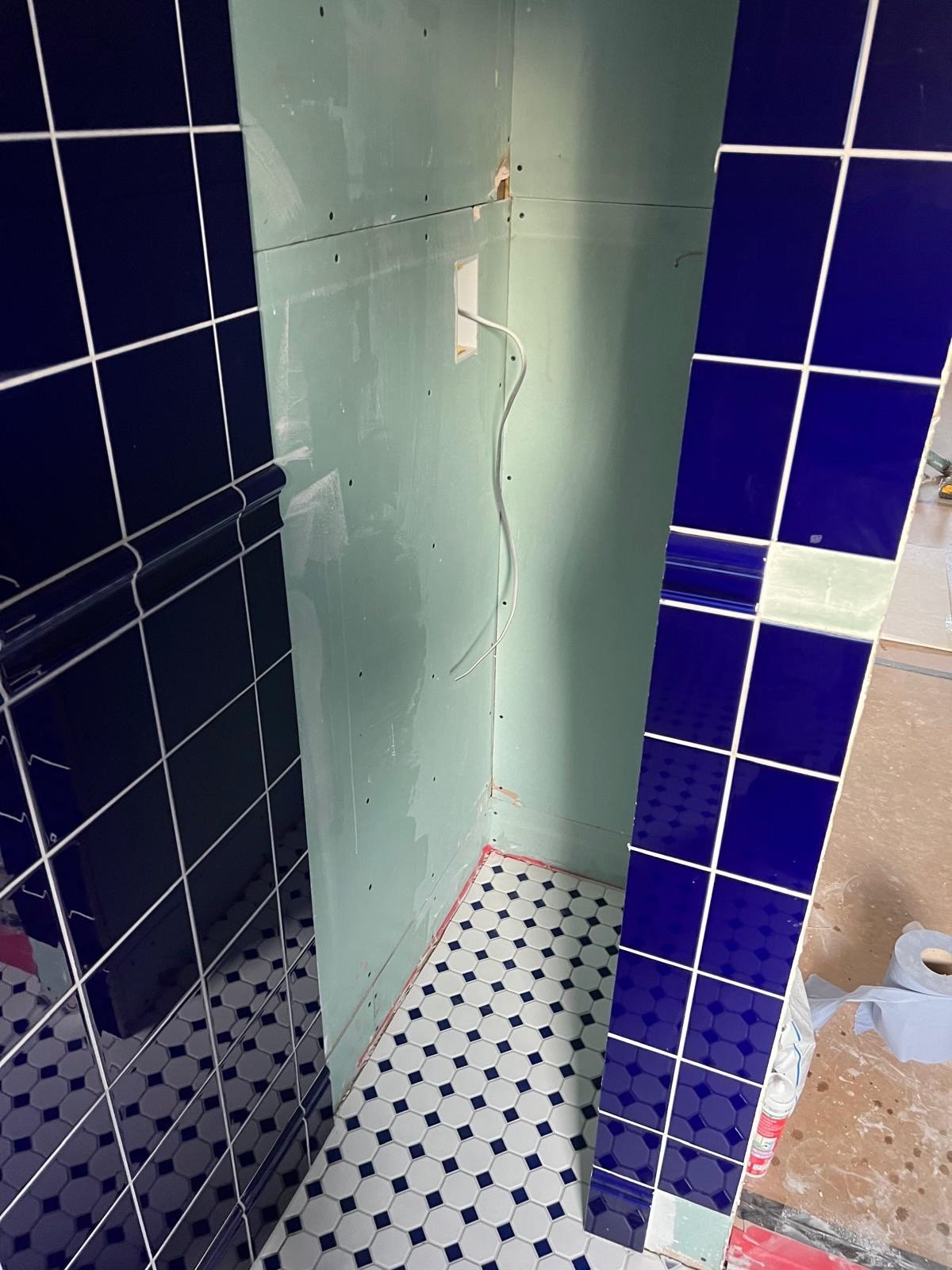 Inside a partially tiled bathroom under renovation with black and blue tiled walls, green drywall, and a patterned hexagon floor. Exposed electrical wiring is visible.