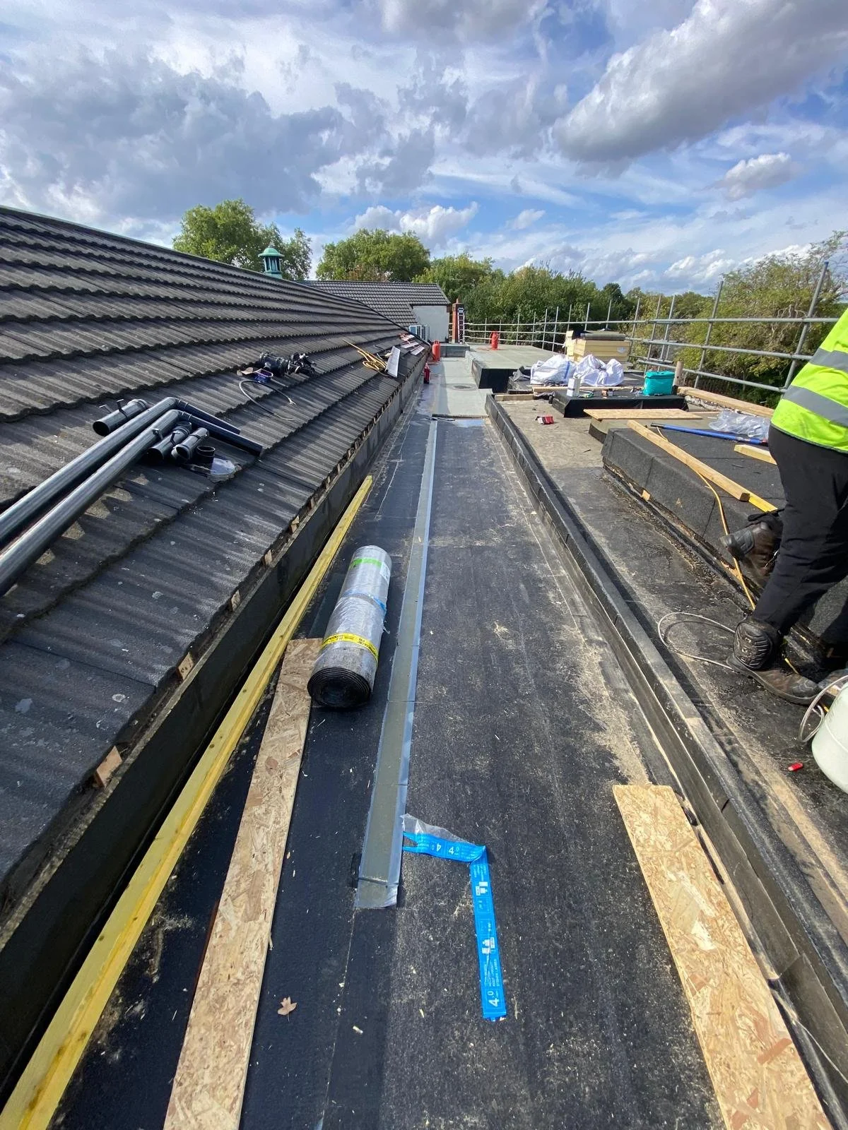 View of a roof renovation site with roofing materials, tools, and construction workers working on the flat roof of a building under a partly cloudy sky.