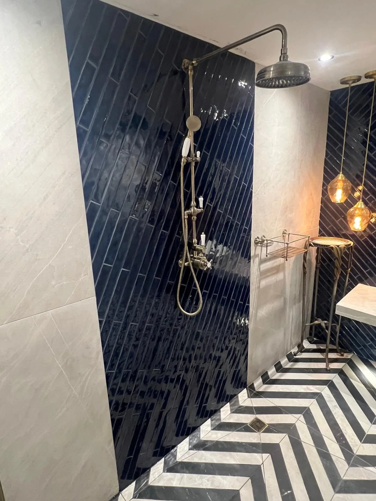 A modern bathroom shower area with black tiled walls, a silver showerhead, and matching fixtures. The floor features a black and white striped chevron pattern. There are pendant lights with warm tones hanging from the ceiling, and a small towel rack 