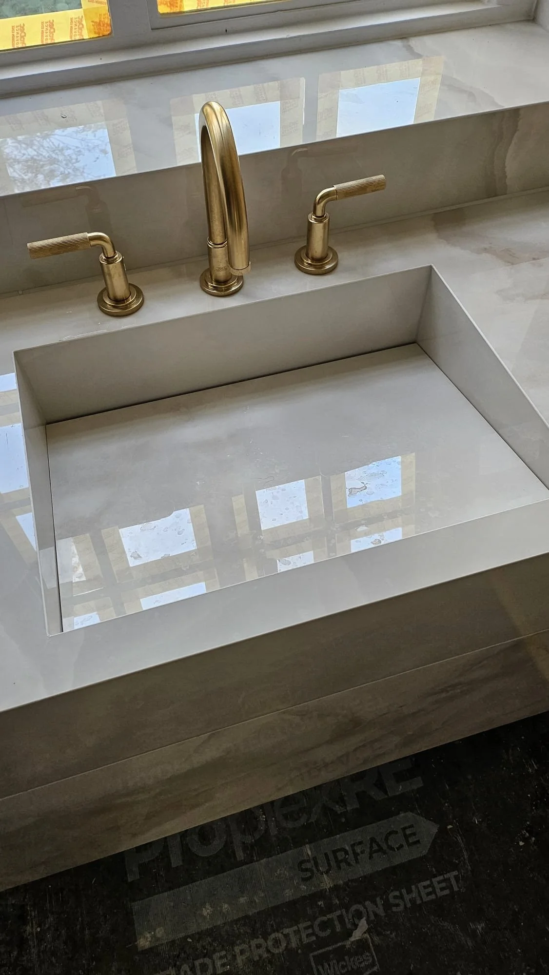 A modern kitchen sink with a gold faucet and handles, installed in a white marble countertop near a window.