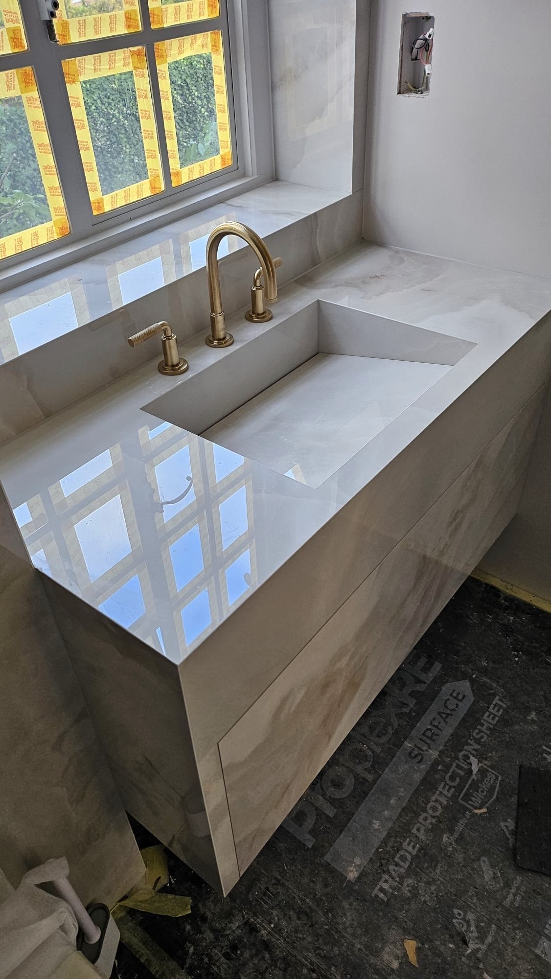 Marble kitchen sink and countertop with a gold faucet next to a window, with construction materials and wiring visible.