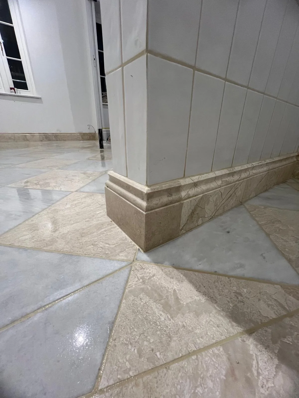 Close-up of a corner with tiled walls and marble flooring, detailing the baseboard transition between the tiles and wall.