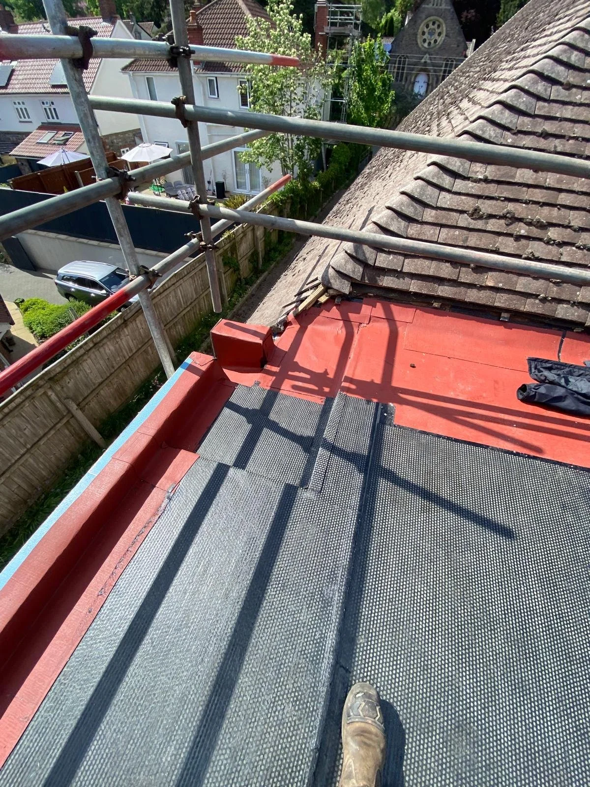 View from a rooftop scaffold showing roof tiles, greenery, neighboring houses, and a person's boot in the corner.