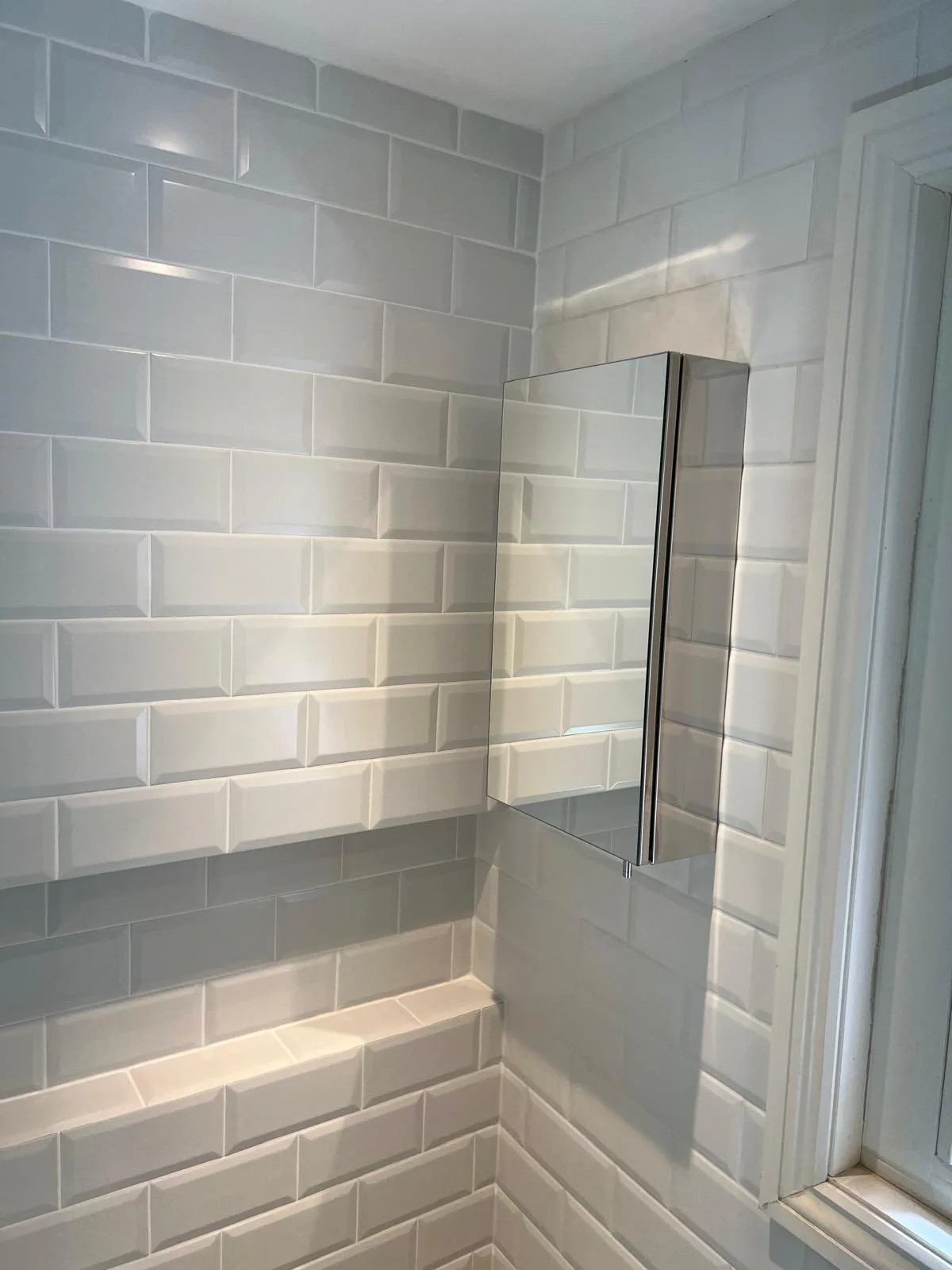 A tiled bathroom wall with a mirror cabinet and a window visible in the corner.