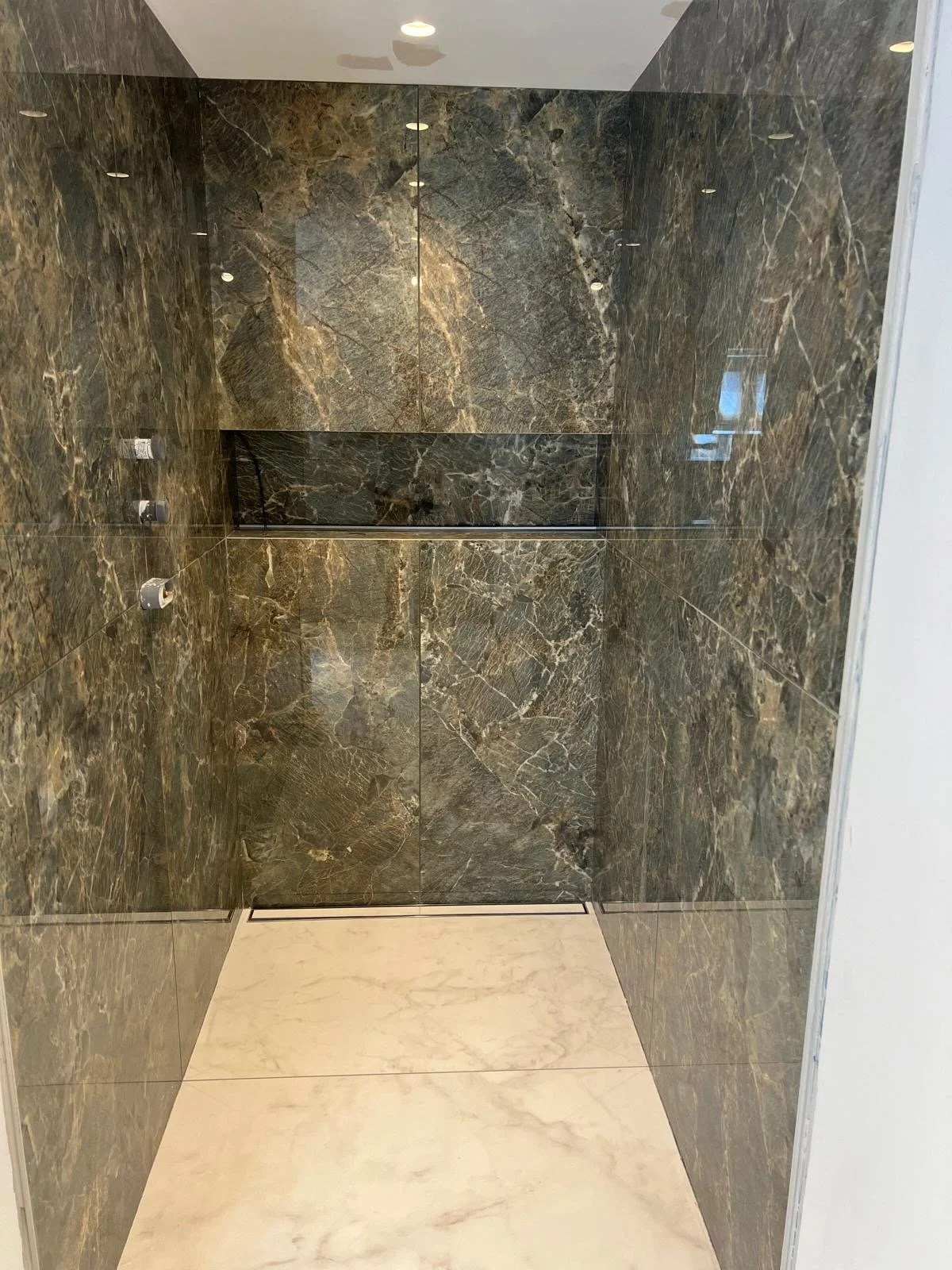 Interior view of a modern elevator with marble walls and floor, with ceiling lights reflected on the shiny surfaces.