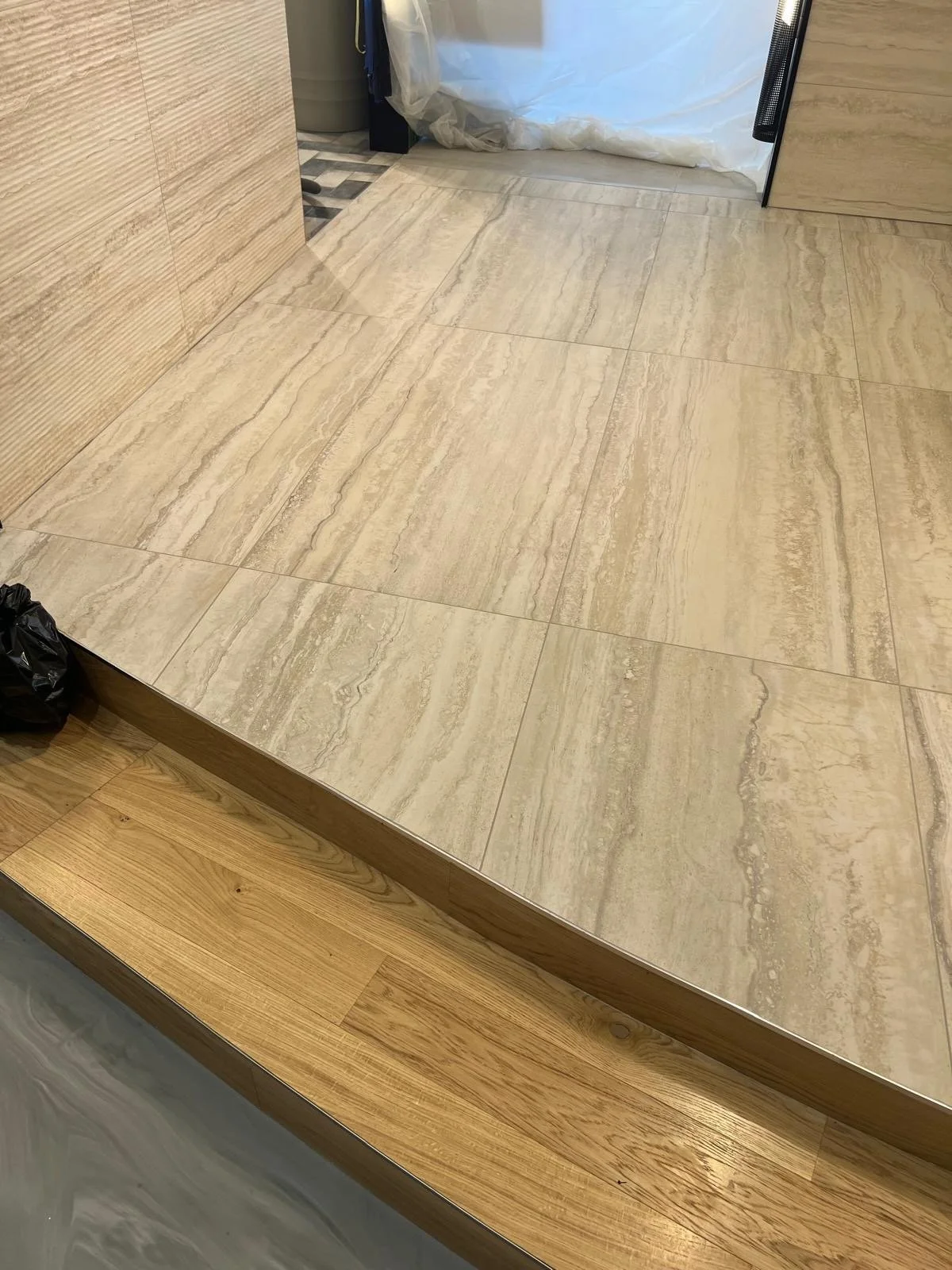 Photo of beige and light brown tiled flooring and a wooden step, with part of a small black trash bag visible in the lower left corner. An area covered with a white sheet or plastic in the background.