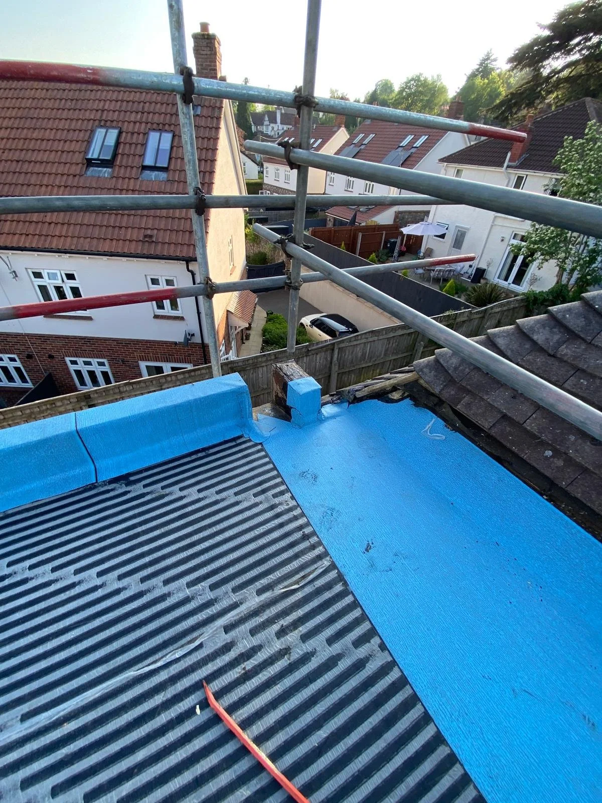 Construction on a roof with blue waterproofing membrane and scaffolding, with neighboring houses visible in the background.