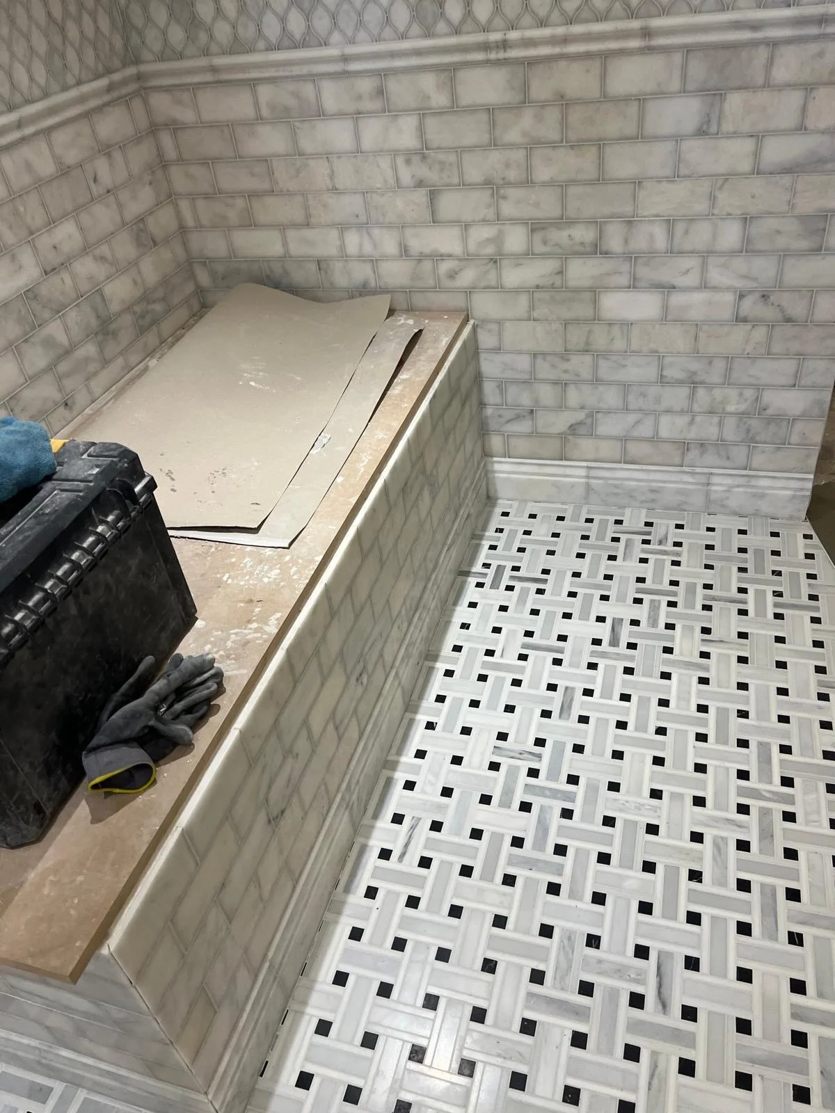 A partially tiled bathroom corner showing marble tiles on the wall and geometric black and white patterned tiles on the floor. There are construction tools and gloves on the countertop.