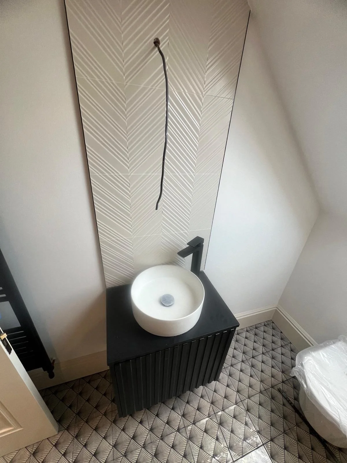 A bathroom with geometric patterned floor tiles, a black vanity with a white round vessel sink, a wall-mounted faucet, and a decorative tile wall behind the sink.