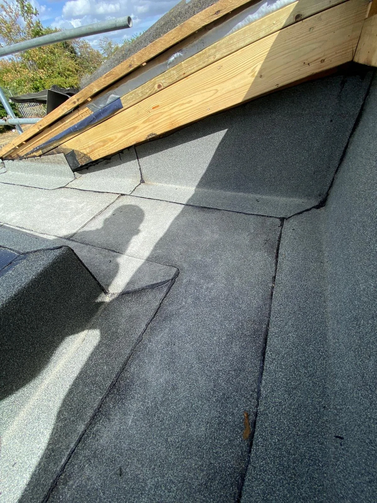 Close-up of a roof with roofing shingles, wooden underlayment, and scaffolding, with some trees and construction materials in the background.