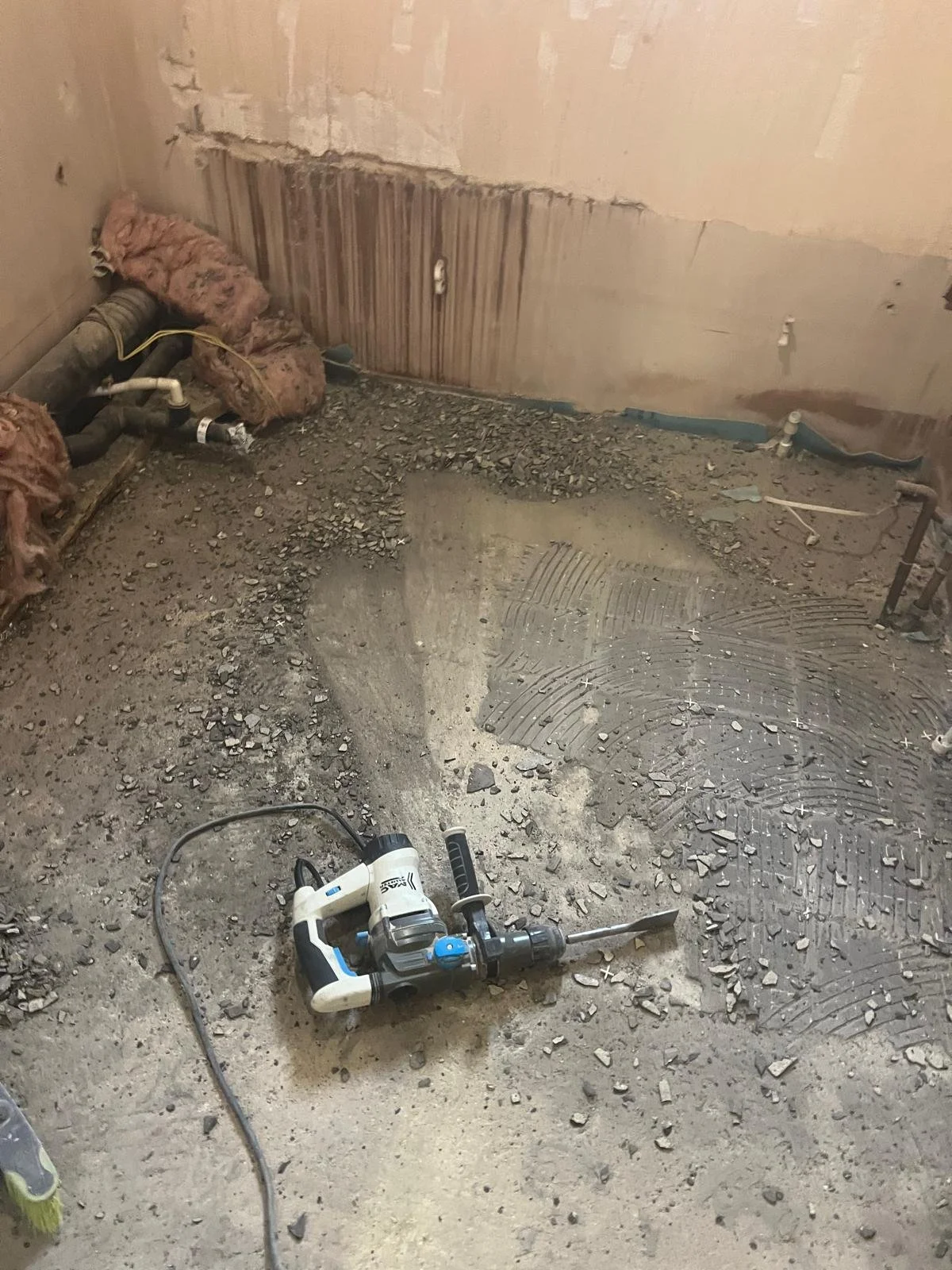 Construction site with an exposed floor, debris, insulation materials, pipes, and a power tool on the ground.