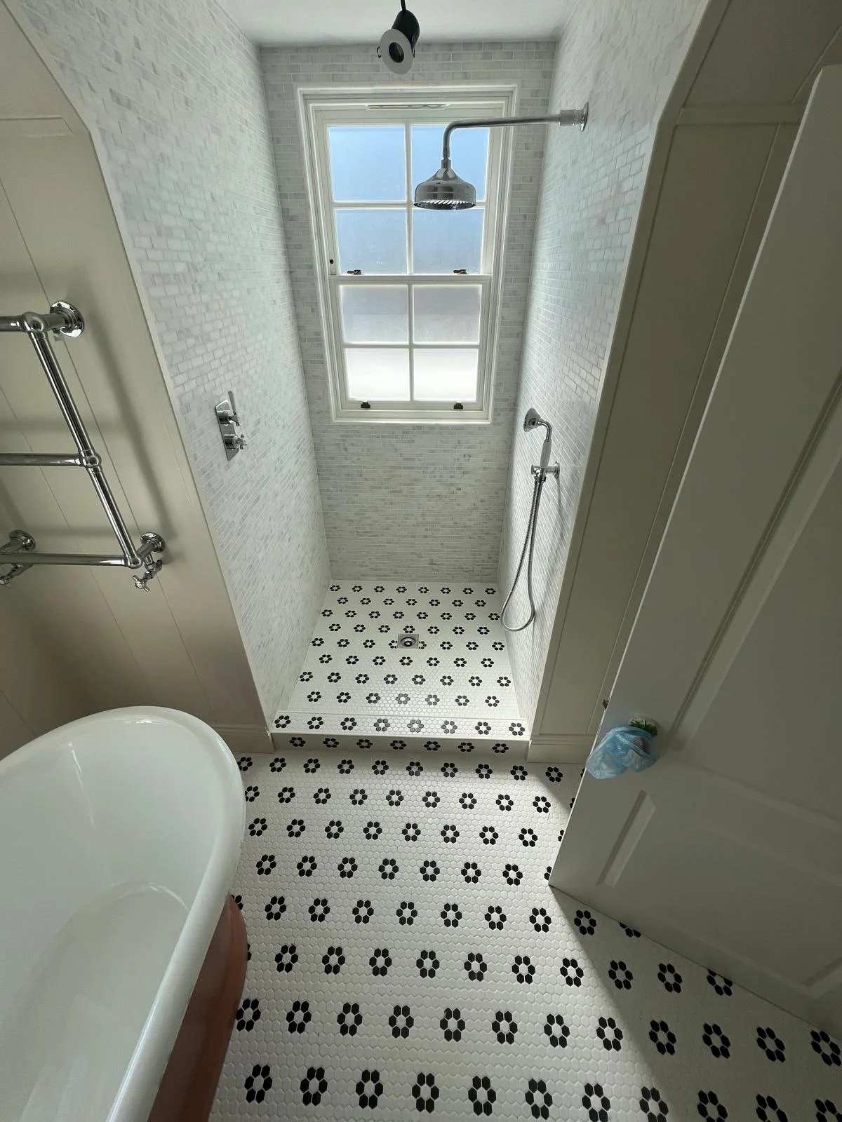Modern bathroom with a walk-in shower containing black and white floral patterned tile floor, white brick walls, and a large window. There is a bathtub partially visible in the foreground, and a ceiling-mounted shower head with an overhead light fixt