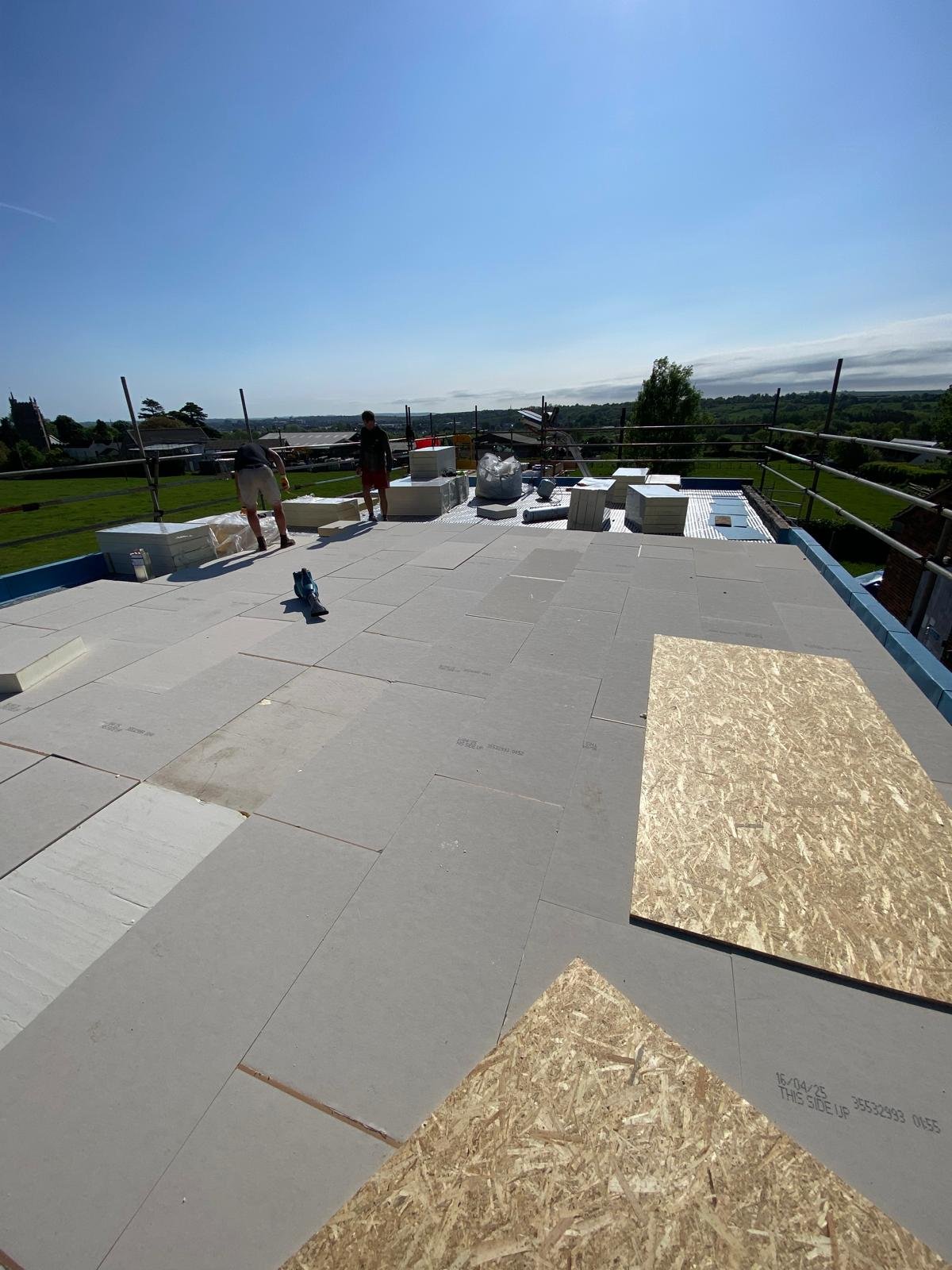 Construction workers installing large light-colored tiles on a flat rooftop outside under a clear blue sky.