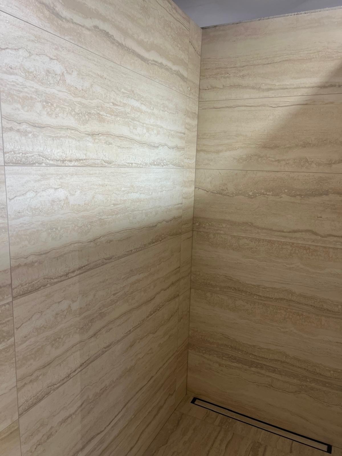 Bathroom corner with beige marble tiles on walls and floor, featuring horizontal natural striations and a linear drainage grate on the floor.