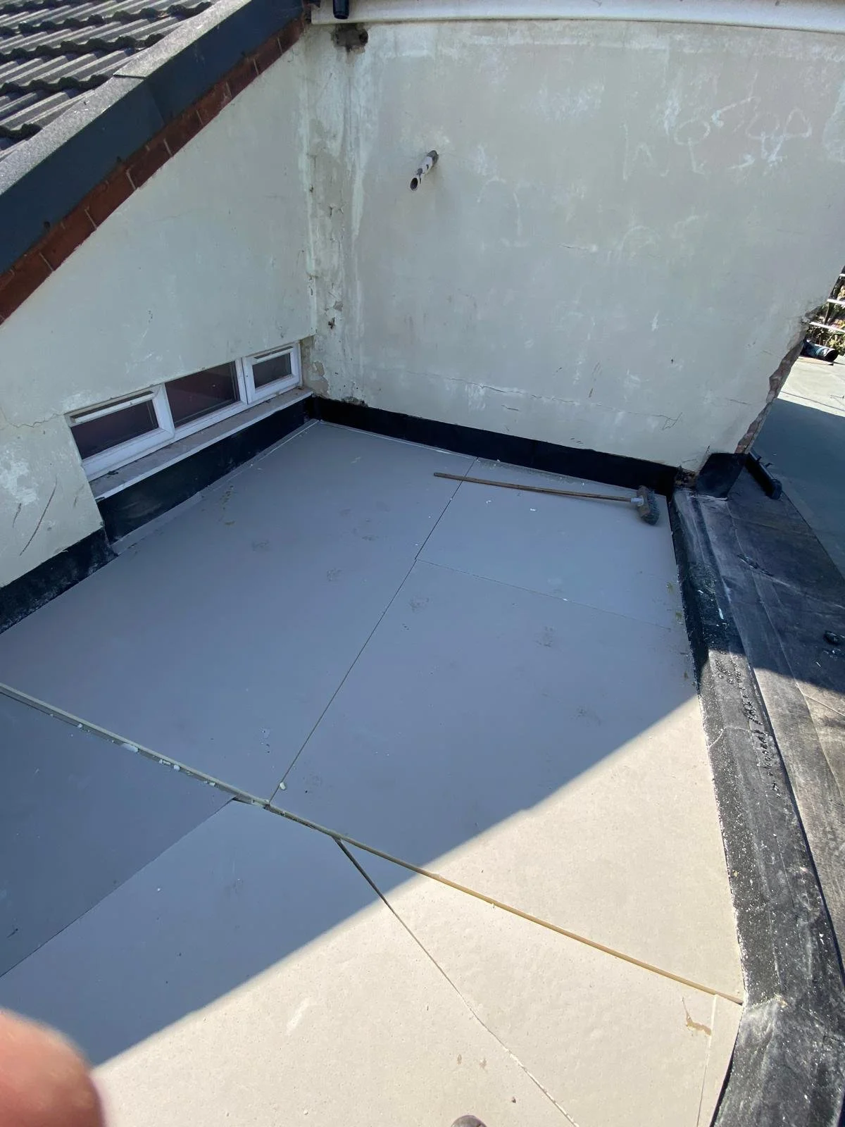 A rooftop with a newly installed flat gray waterproof membrane, a small window, and a white wall with exposed and peeling paint.
