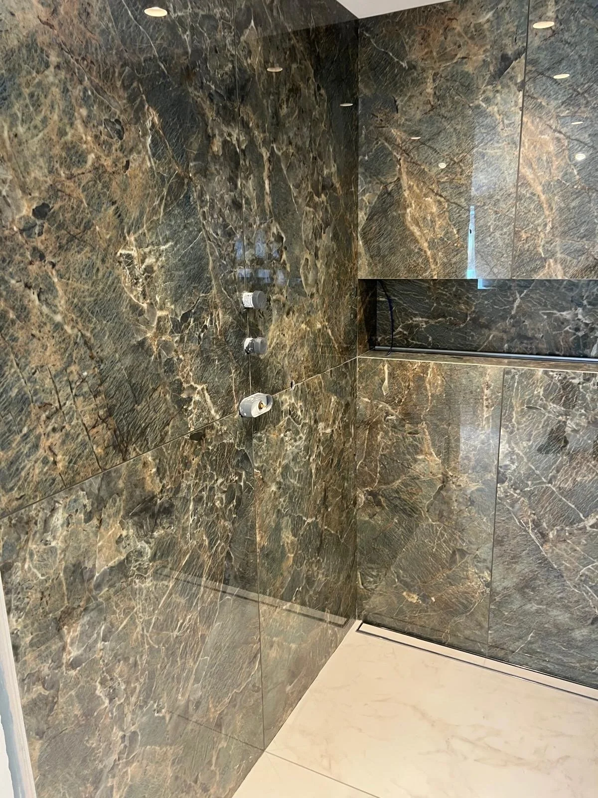 A modern shower with dark brown and gray marble walls and a white marble floor. Four showerheads are mounted on the wall, and a mirrored panel reflects part of the shower area.