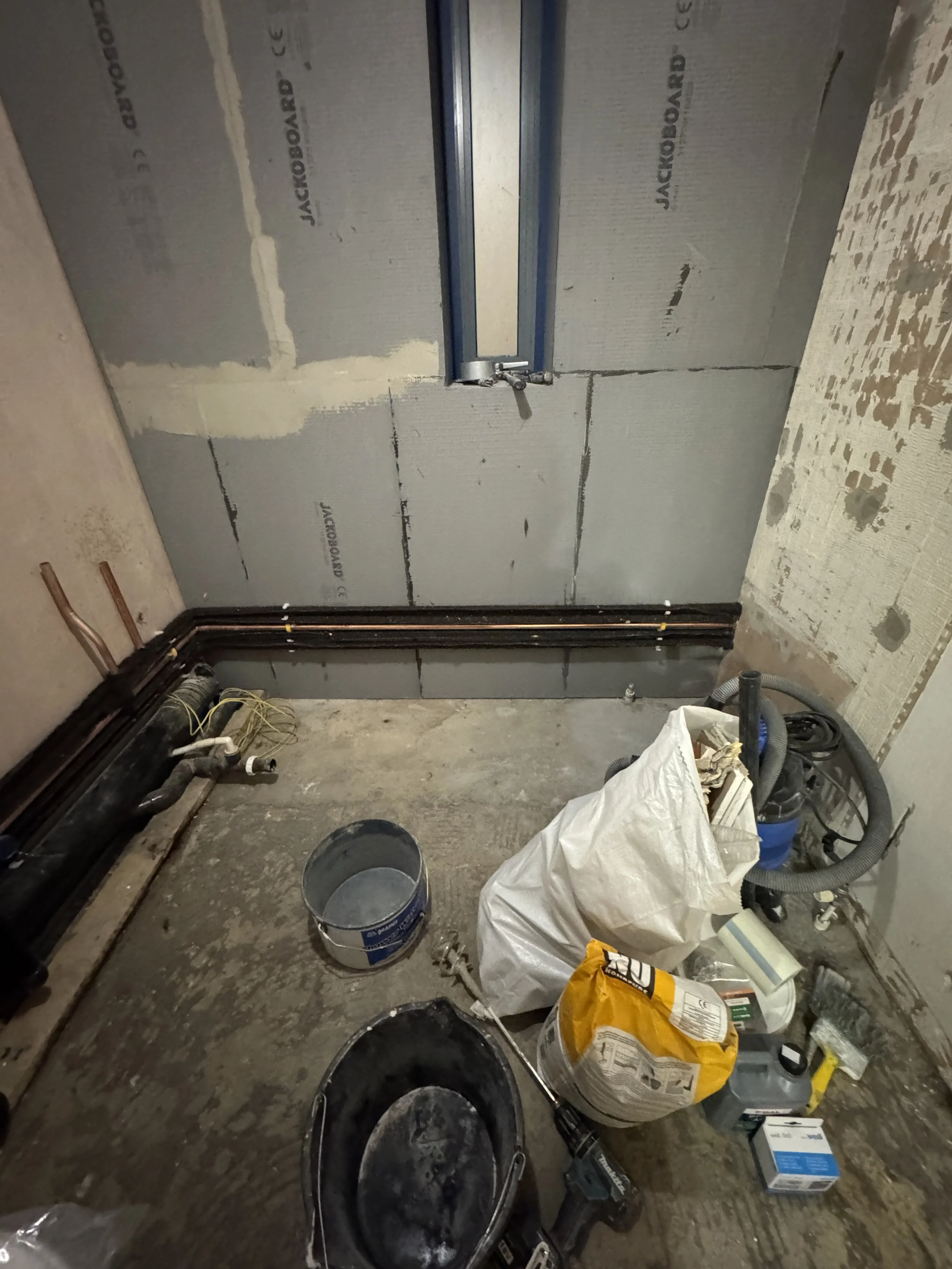 Construction site with tools, pipes, a bucket, a bag of concrete, and an unfinished wall with insulation panels.
