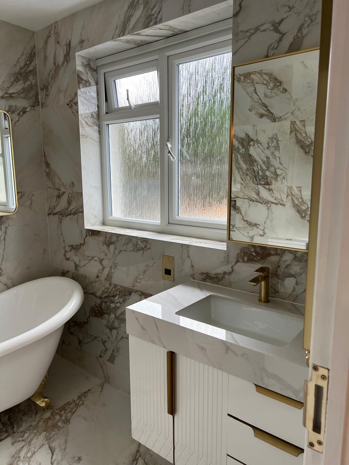 A bathroom with marble walls and floor, a white bathtub, a small marble sink with gold fixtures, a frosted window, and a mirrored cabinet with a gold frame.
