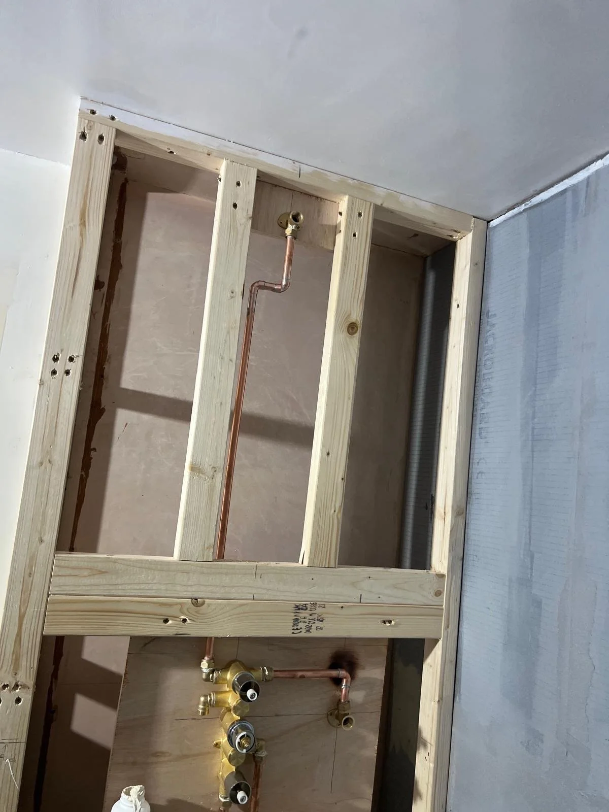 Construction framing for a wall with plumbing and water shut-off valves installed, in an unfinished room with drywall partially installed.