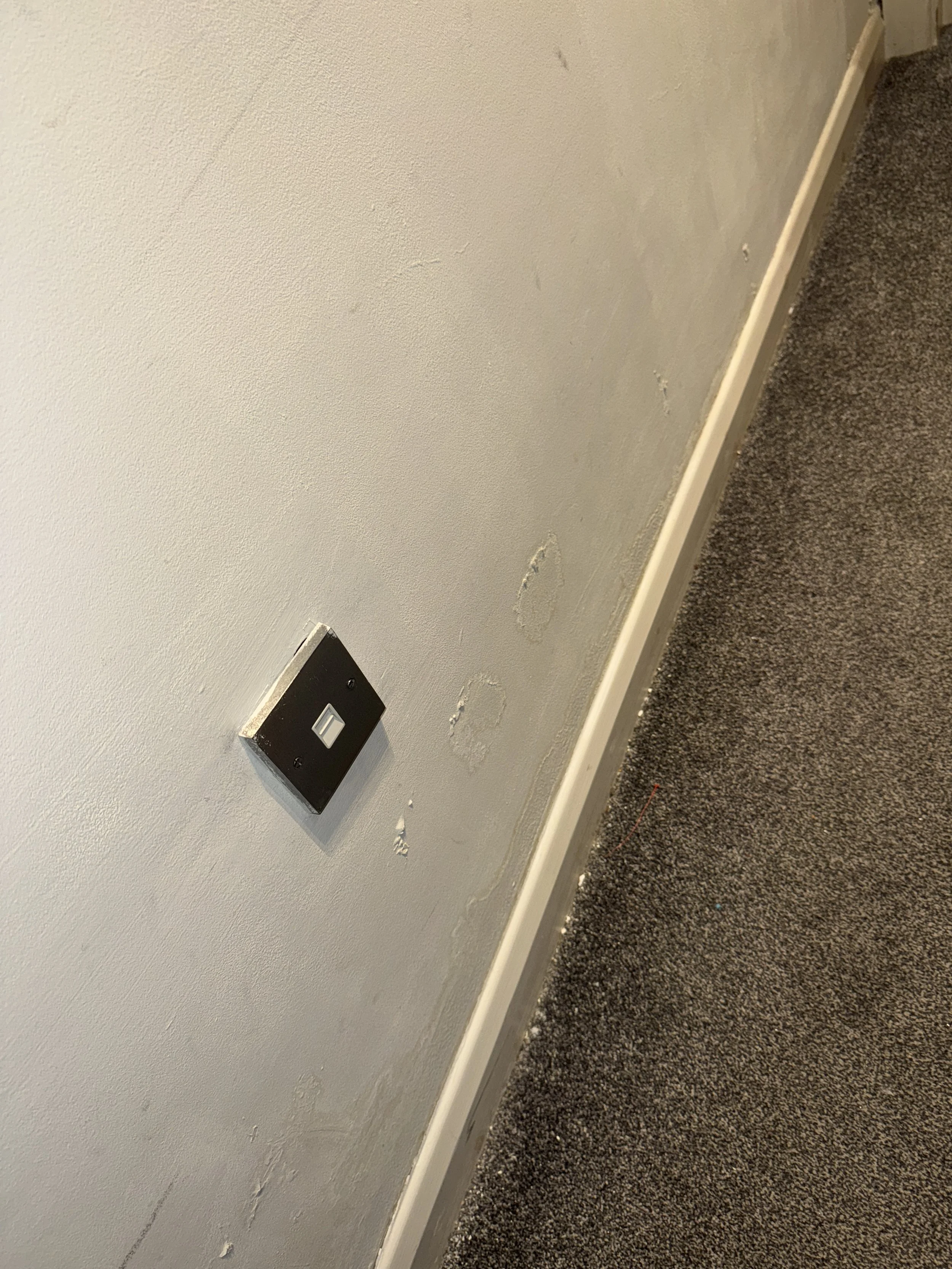 A wall with a black electrical outlet plate, removed patches of paint, and a gray carpeted floor.