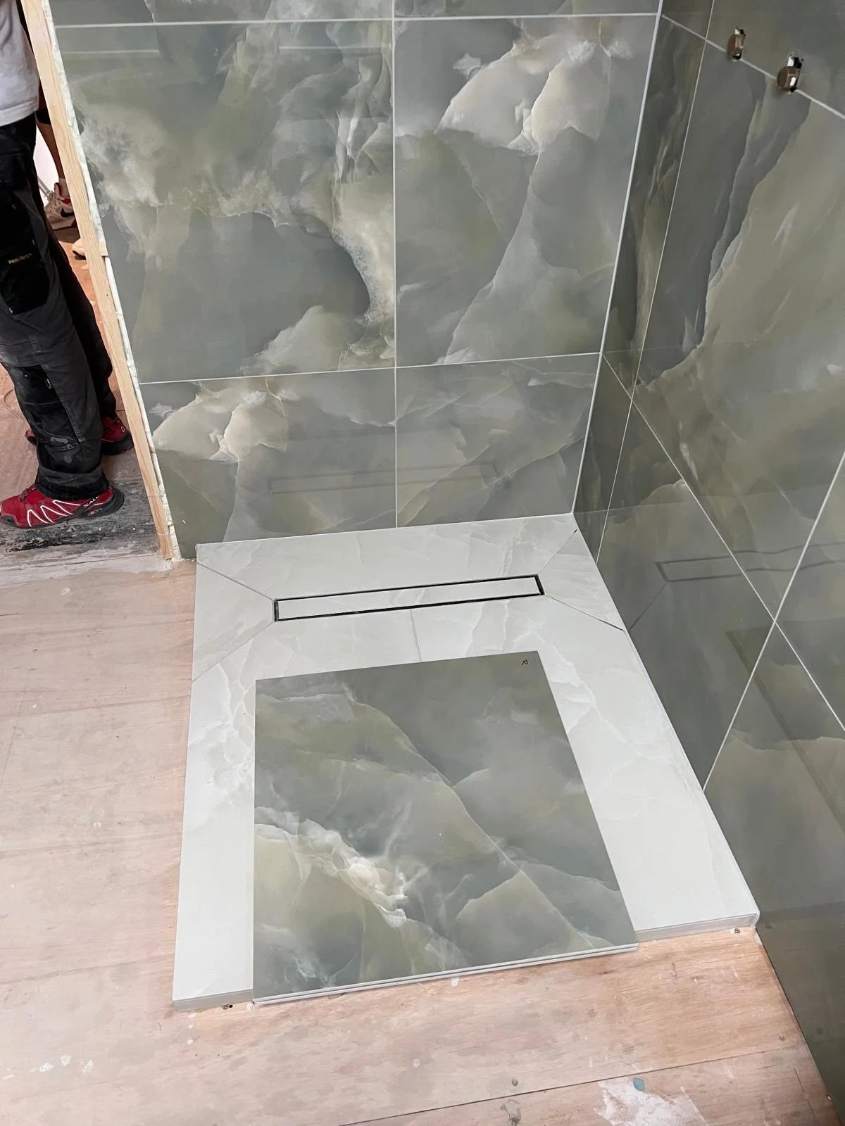 A shower area with large marble-like tiles on the walls and a smaller marble-like tile on the shower floor. There is a linear drain on the shower floor and a person standing nearby wearing black pants and red shoes.