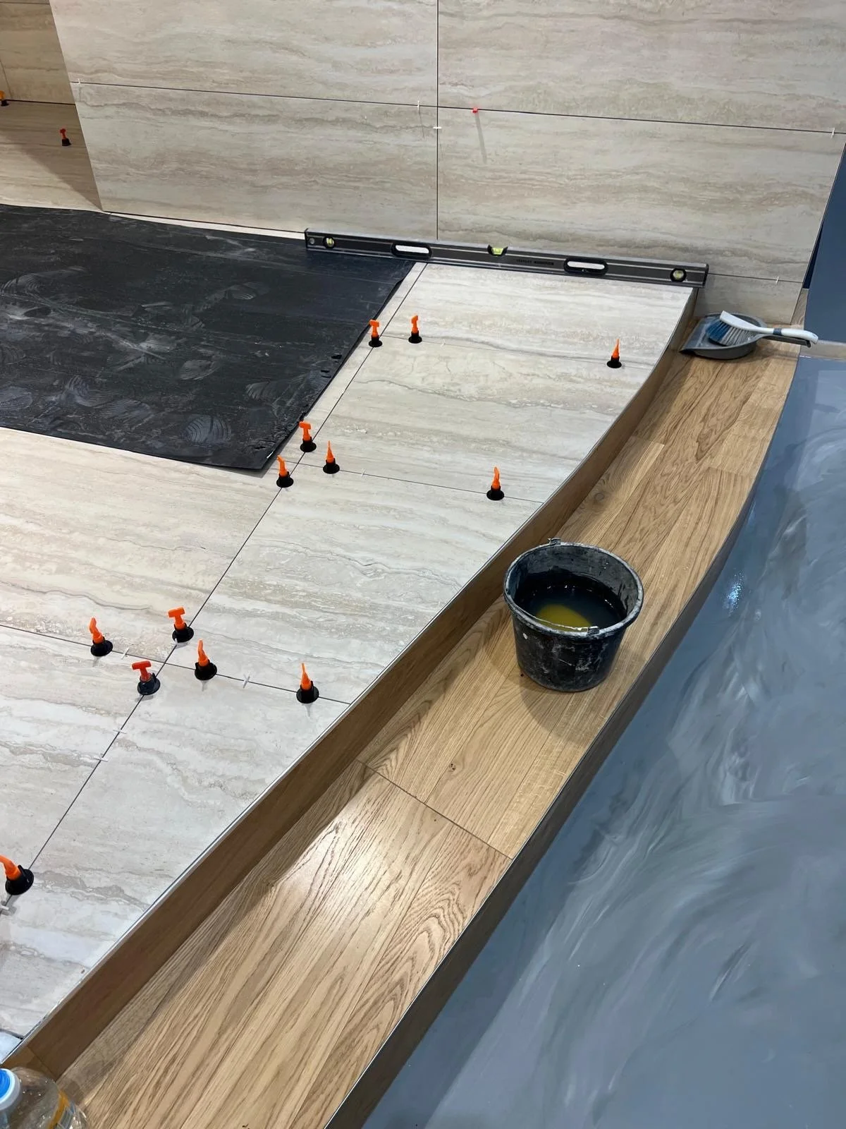 A construction scene showing a partially tiled wall with large tiles, a level tool, plastic tile spacers, a bucket with adhesive or grout, and a small sponge brush on a wooden surface. The wall tiles are being aligned and spaced with a string guide.