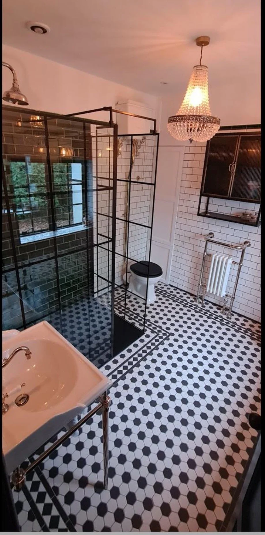 A bathroom with black and white patterned tile flooring, a white brick wall, a black grid shower enclosure, a white sink with chrome fixtures, a black toilet with a black seat, a vintage-style radiator, a black wall cabinet, and a ceiling chandelier.