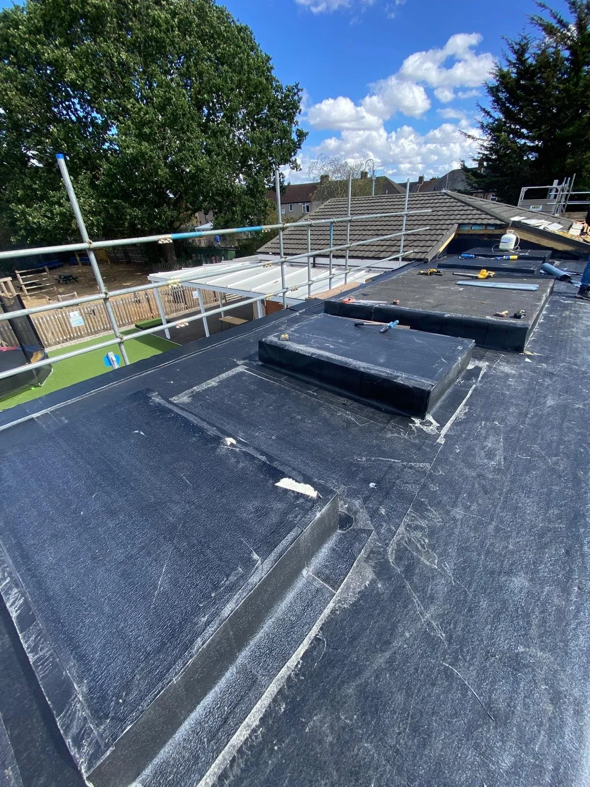 Roof under construction with black roofing material, vents, and tools, surrounded by scaffolding, with neighboring homes, trees, and a blue sky with clouds in the background.