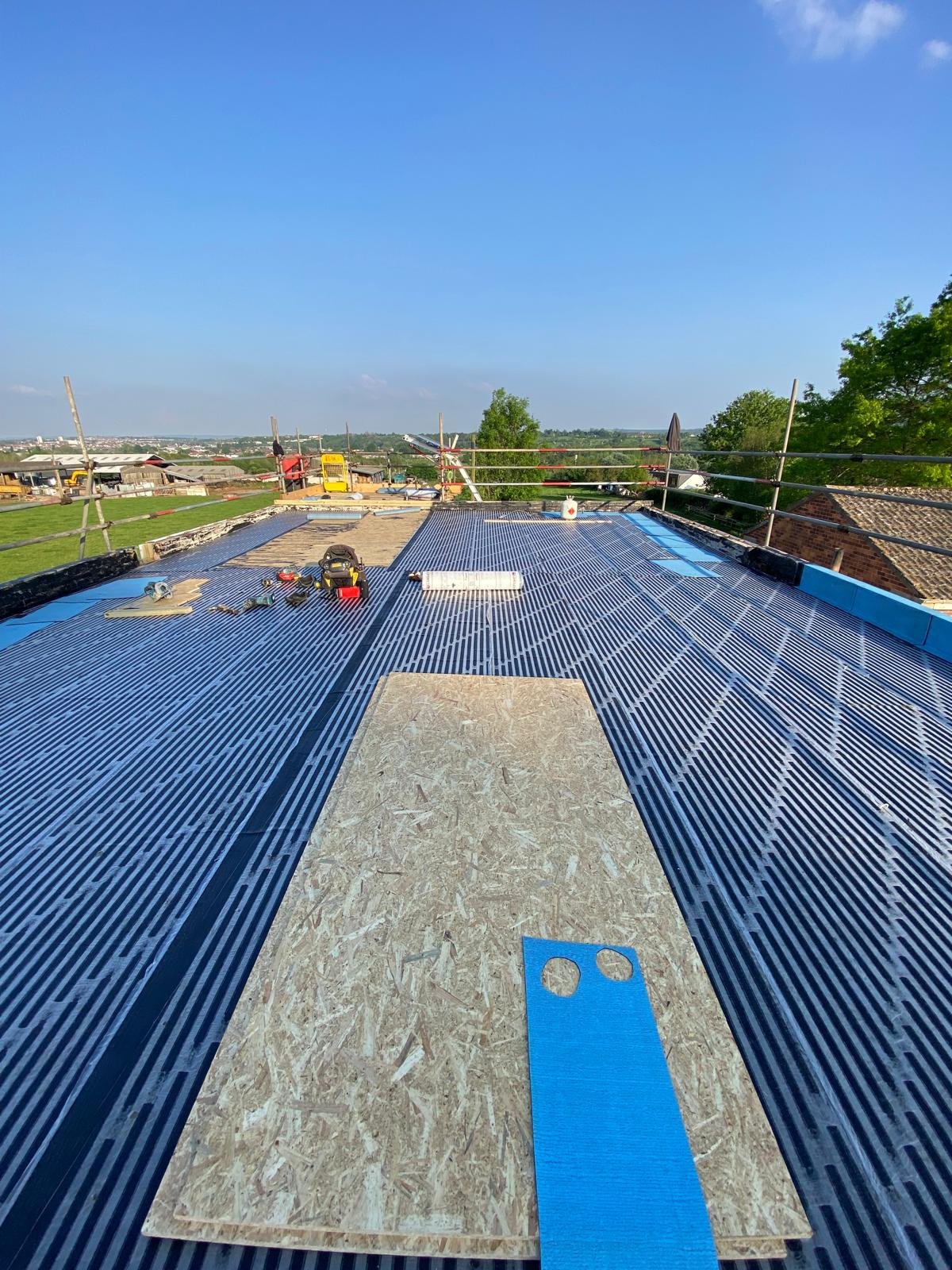 Roof construction with metal roofing panels, plywood boards, tools, and equipment on a sunny day.