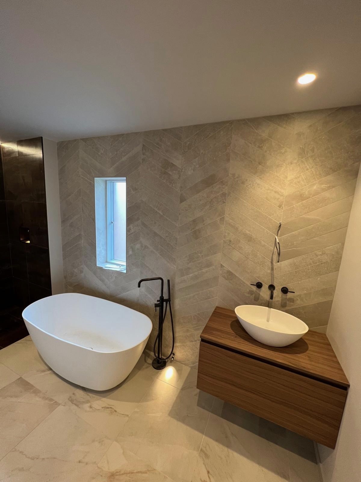 Modern bathroom with a freestanding bathtub, a narrow window, a wooden vanity with a vessel sink, and textured beige wall tiles.