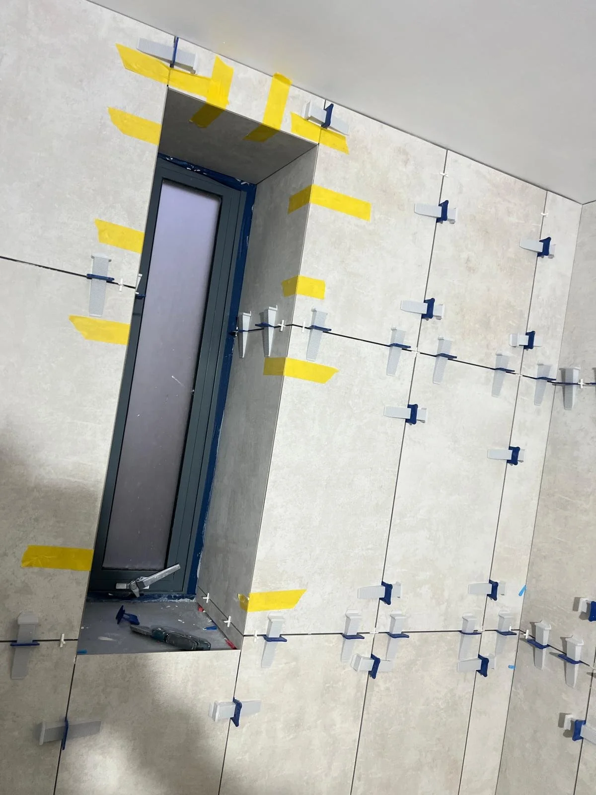 Bathroom wall with tile installation in progress, tiles are being secured with spacers and masking tape around the window frame.