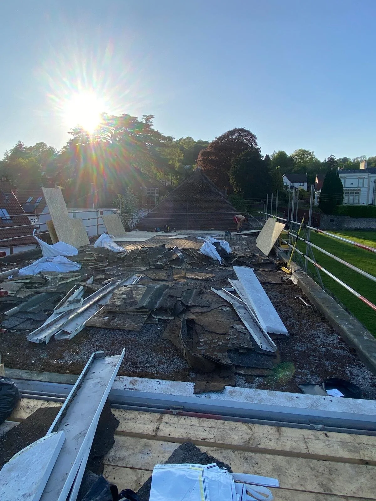 Roof under construction with scattered tiles and construction materials, workers in background, sunny clear sky, trees and houses in the distance.