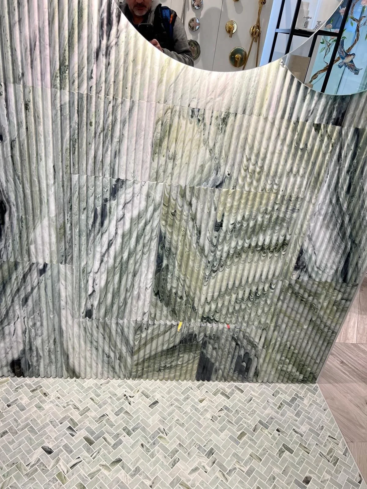 Close-up of a marble partition with a curved mirror above, showing the reflection of a person taking the photo and decorative items on the wall, including wall-mounted showerheads and a bird illustration.