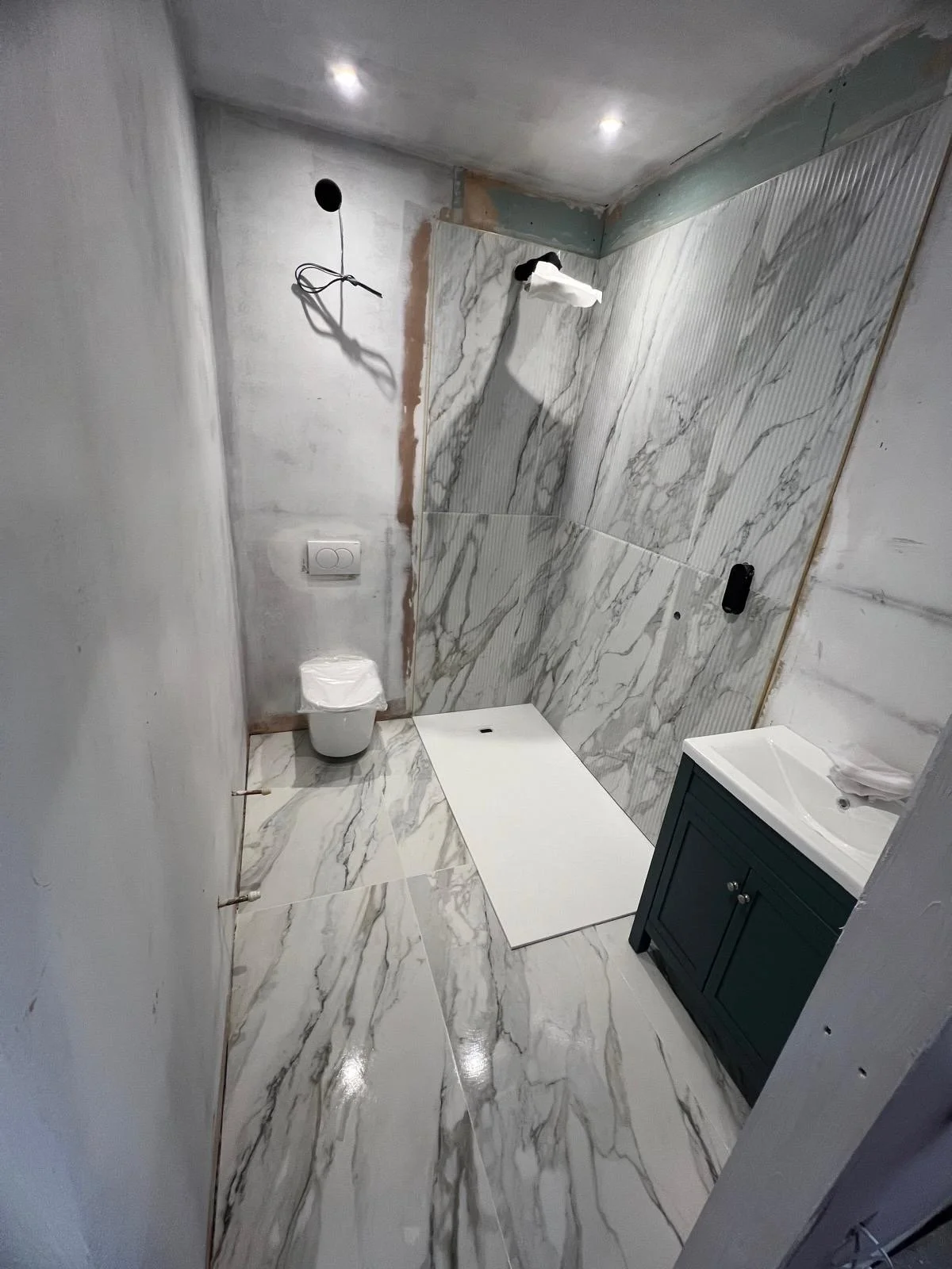 Bathroom under construction with marble tiles on walls and floor, a toilet, a small sink with a cabinet, and an unfinished shower area.