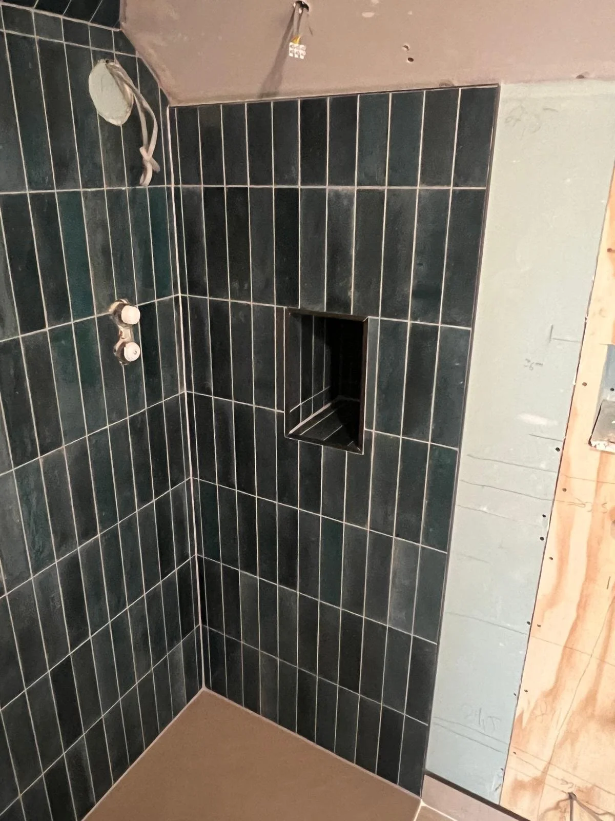 A shower area under construction with black tile walls, a built-in shelf, and exposed plumbing for a shower head and controls.