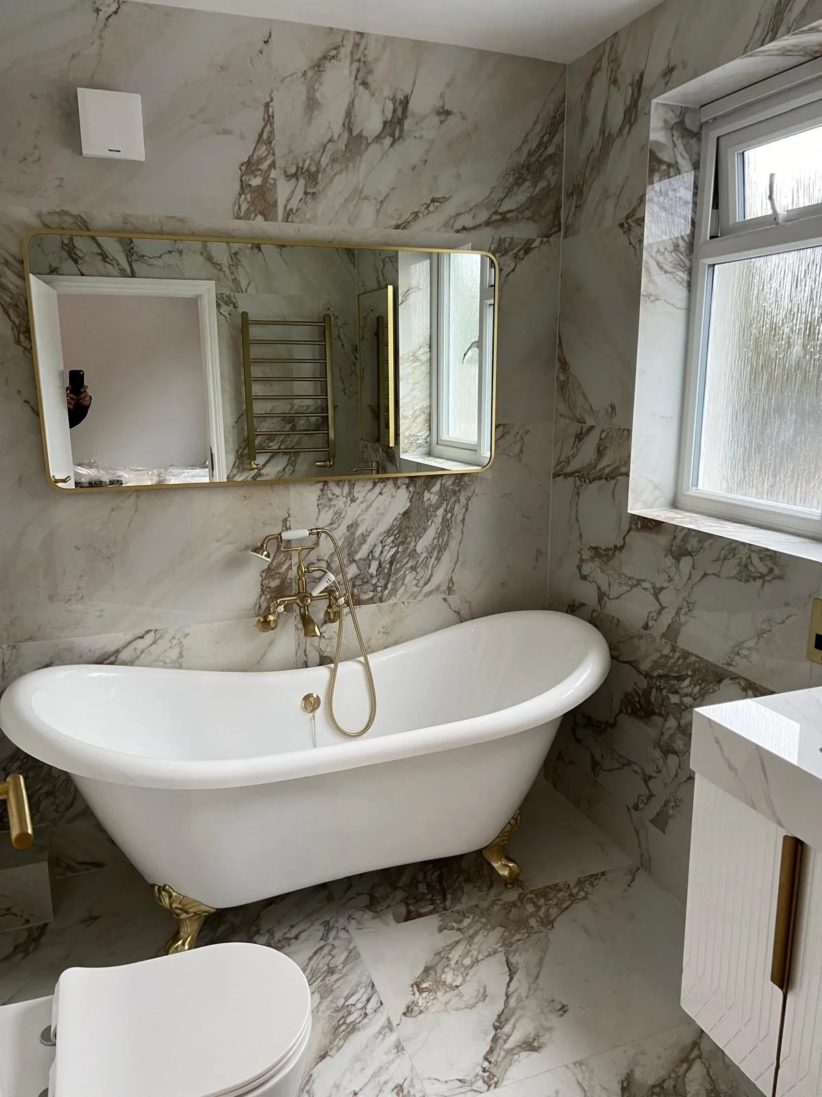 A bathroom featuring a vintage clawfoot bathtub with gold accents, marble walls and floors, a large rectangular mirror, a window, and a small white cabinet.