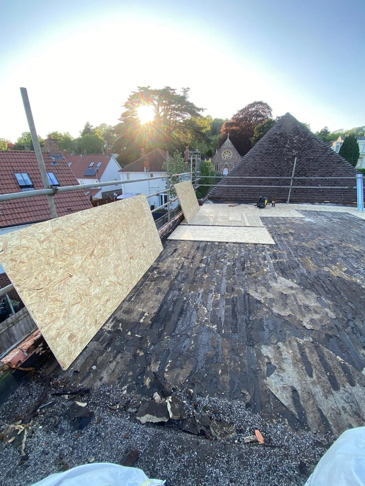View of a rooftop under construction with scaffolding, plywood boards, and tools, overlooking a residential neighborhood with rooftops, trees, and the sun setting behind. Rocky surface on the roof indicating ongoing work.