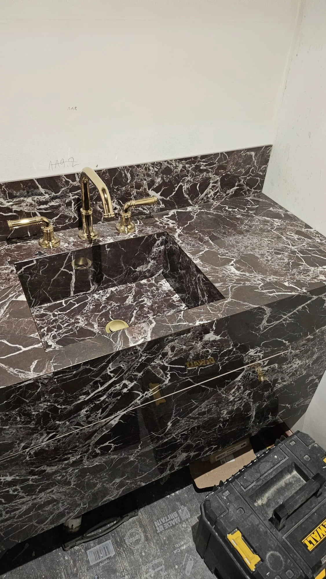A marble bathroom sink with gold-colored faucets and fixtures, in a construction or renovation setting with tools and equipment on the floor.