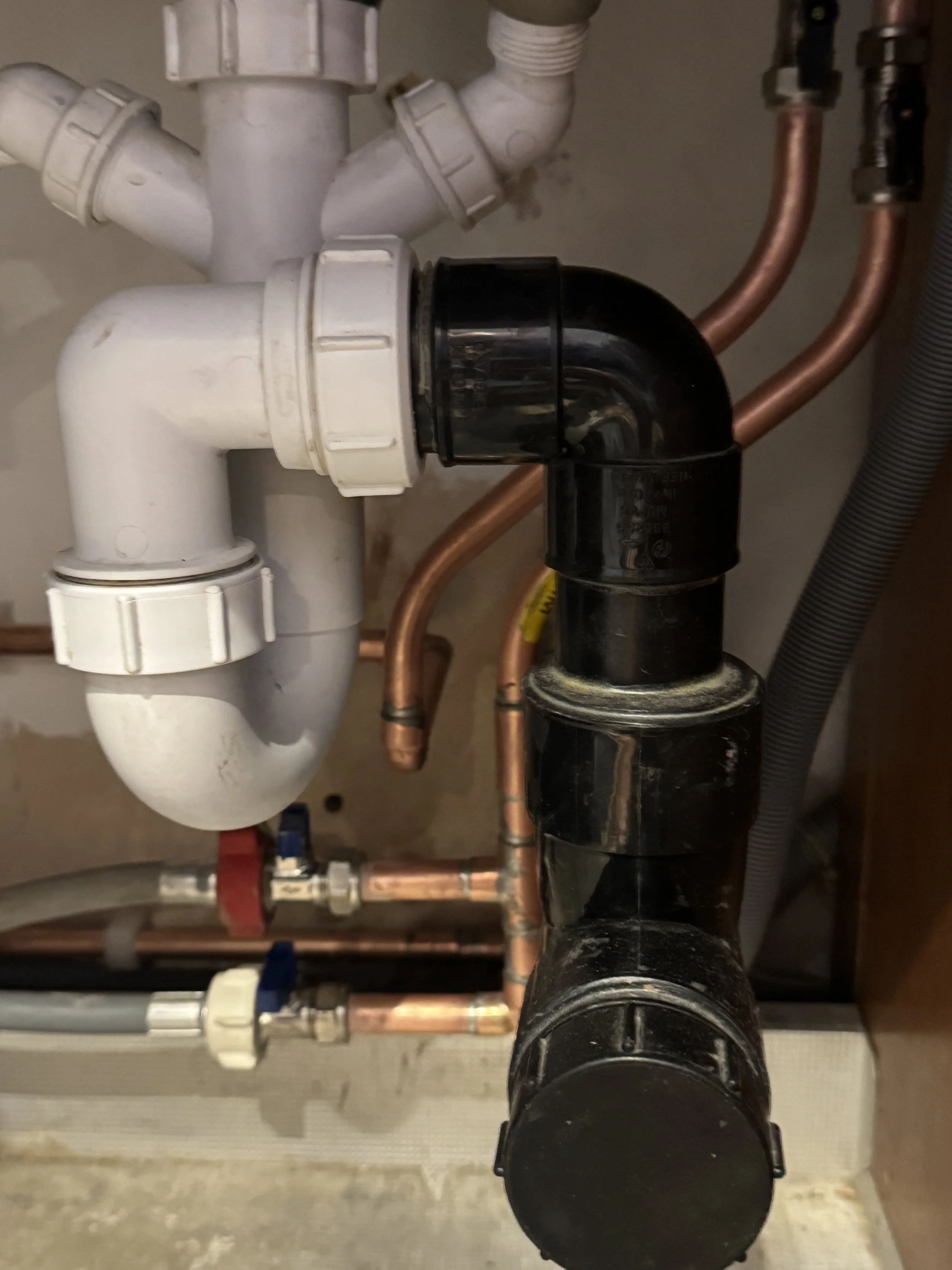 A view of plumbing pipes including white PVC pipes and copper pipes connected to each other, with some fittings and a black pipe in the foreground.