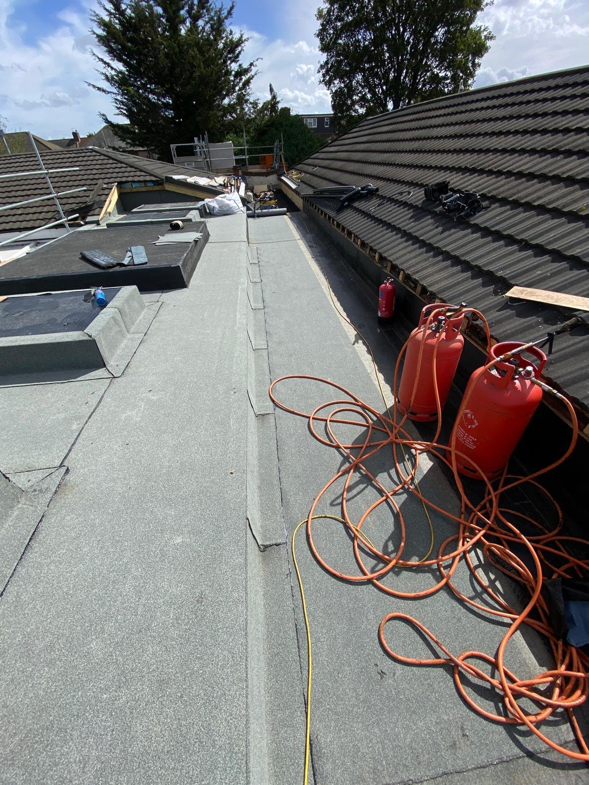 Roof construction site with tools and equipment, including red propane tanks, extension cords, and roofing materials, under partly cloudy skies.