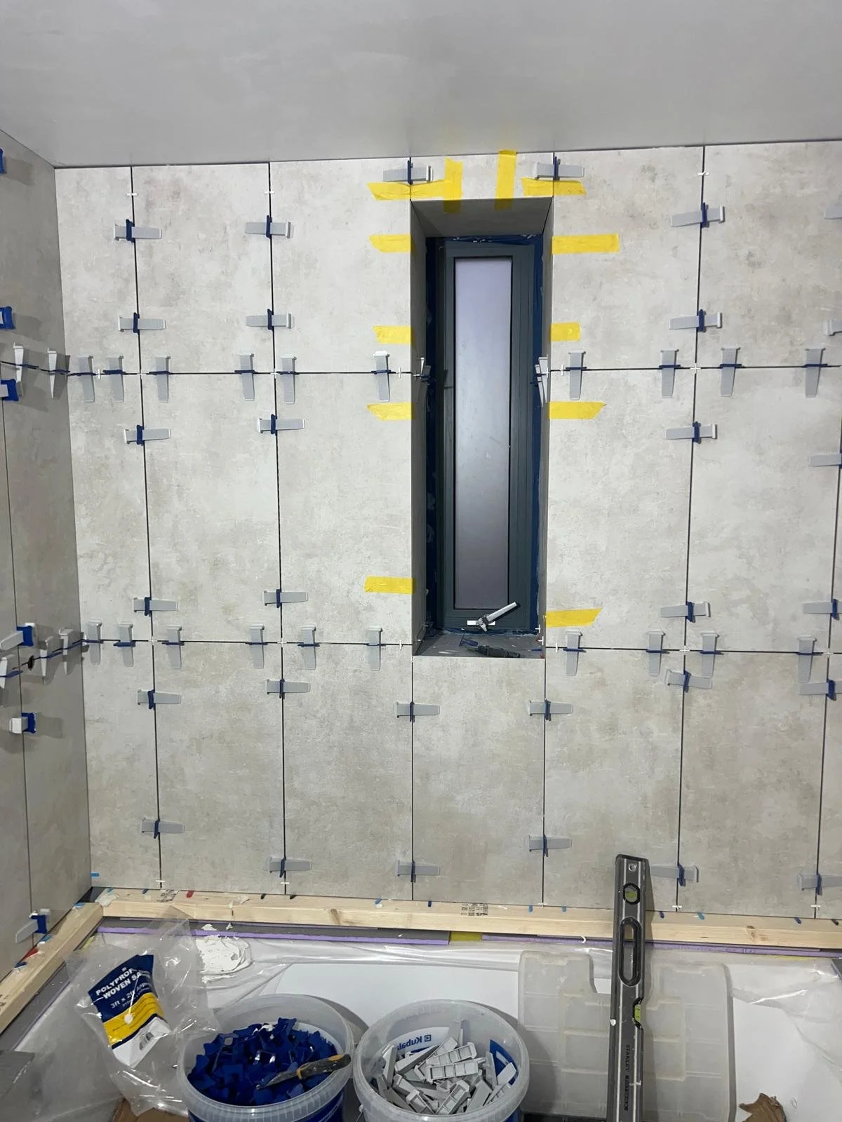 Bathroom wall with tiles being installed, with a small vertical window and construction tools and materials in the foreground.