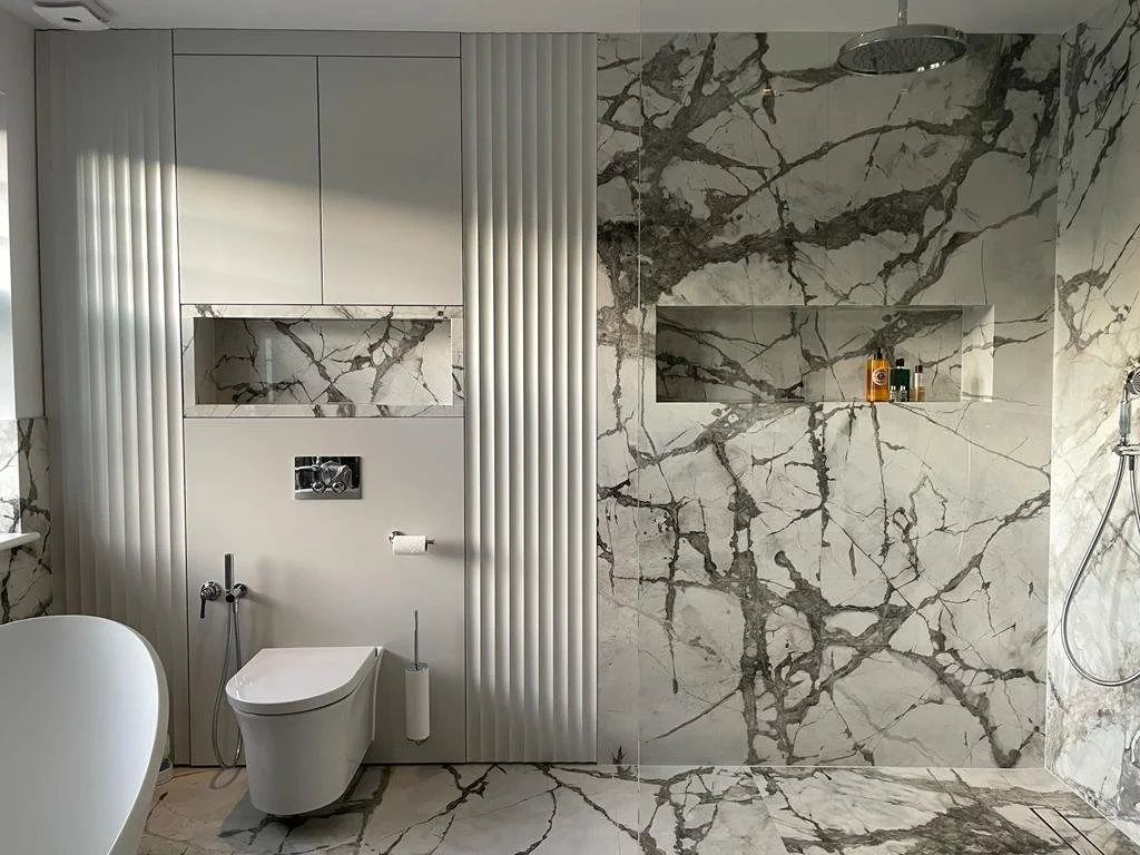 Modern bathroom with white walls, marble tile walls with dark gray veining, a built-in shelf in the wall, a wall-mounted toilet, a handheld shower head, and a rain shower head.