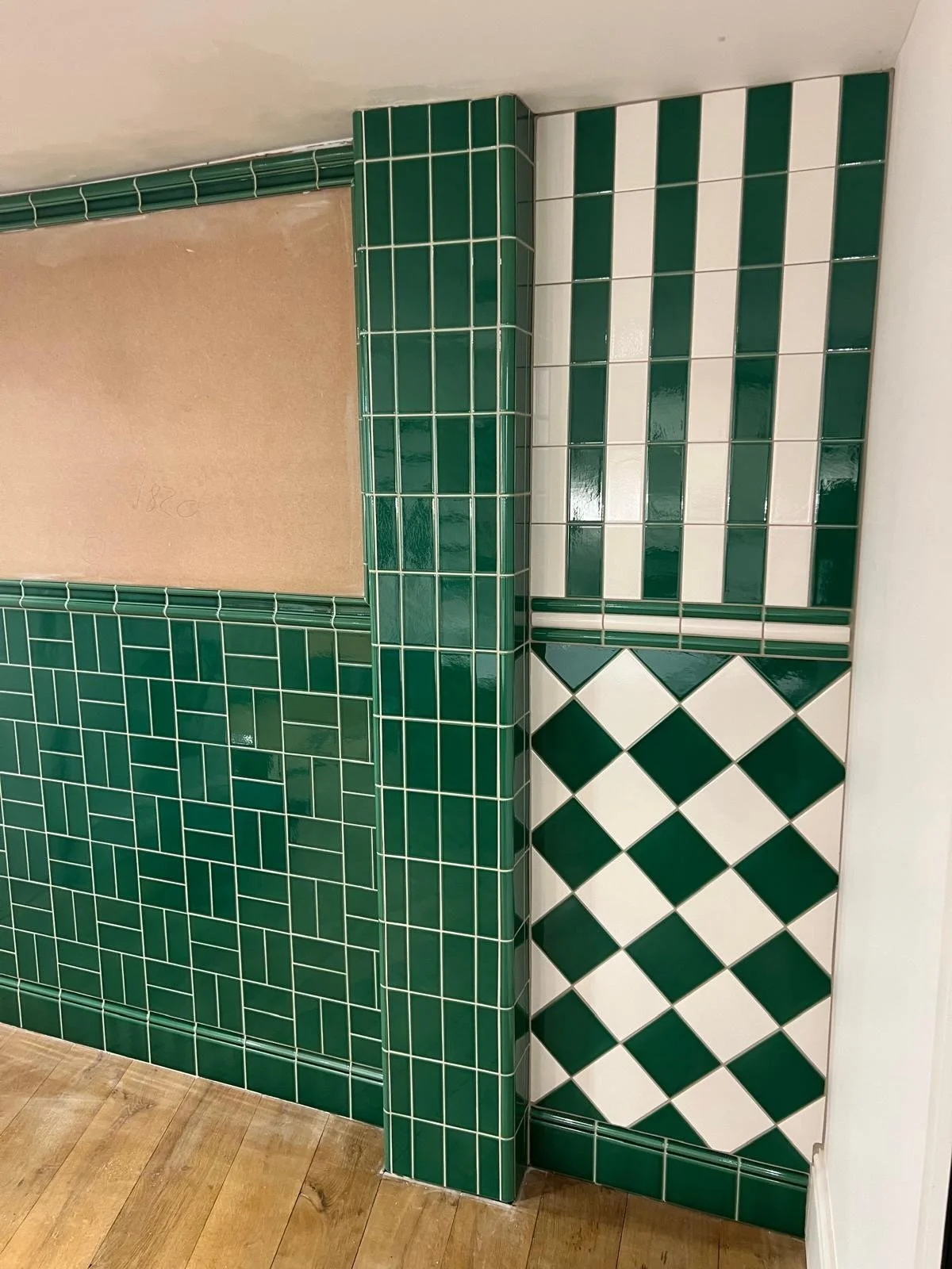 Interior corner with green and white ceramic tiles, checkered pattern on the lower section, rectangular tiles with white grout in middle, and small square tiles in upper section. A wooden floor is visible at the bottom.
