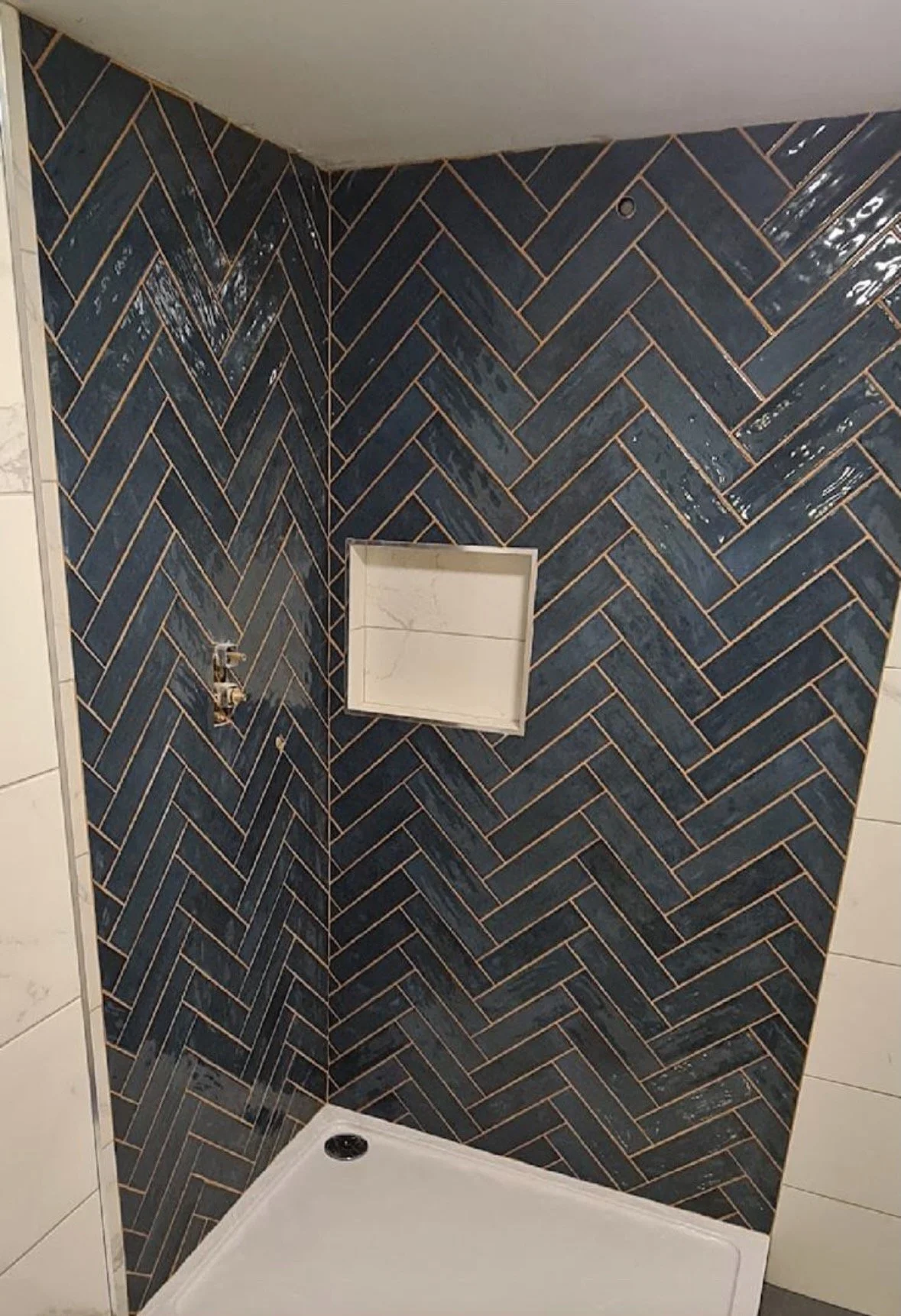 Corner of a shower with dark herringbone tiles, a small unfinished shelf or niche, and visible plumbing fixtures, with light-colored tile on the adjacent wall.