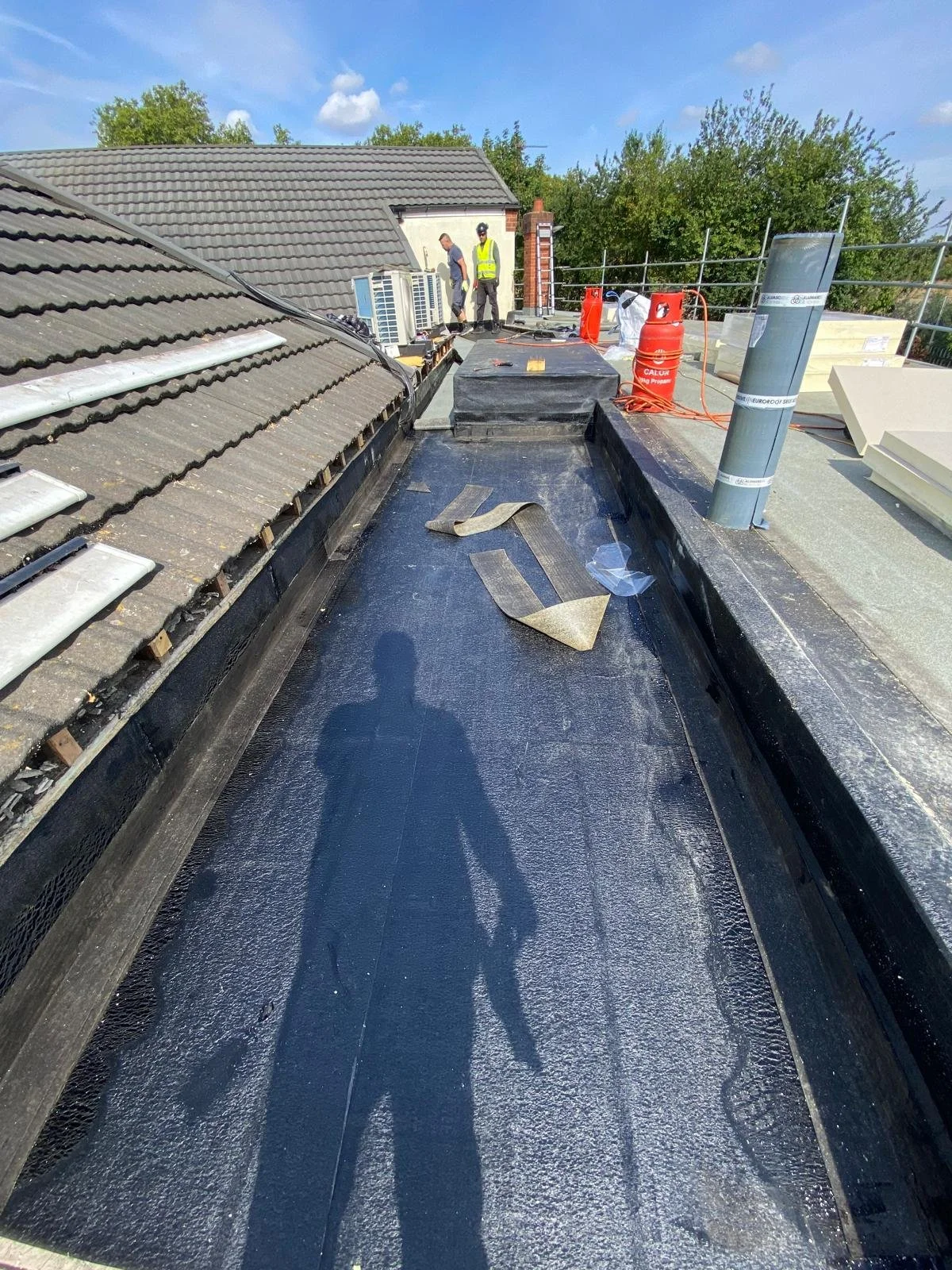 Roof construction workers working on a rooftop with tools and building materials, including a rolled roofing membrane, vent pipe, and propane tank, under a partly cloudy sky.