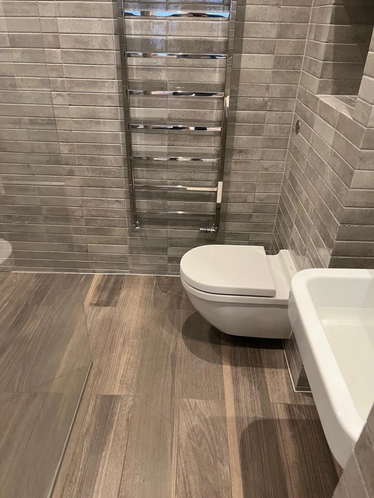 A bathroom with a wall-mounted toilet, a white sink, tile walls in shades of gray, a chrome heated towel rack, and wood-patterned floor tiles.