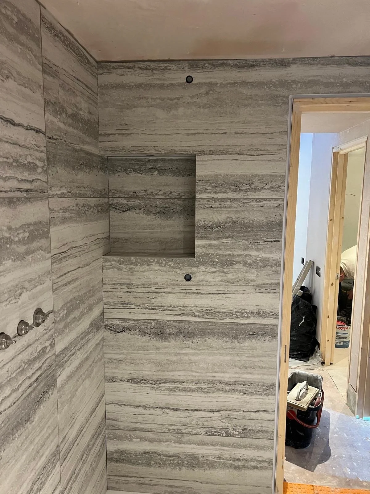 A bathroom under construction with gray stone-look tiles on the wall. There are two small black holes, possibly for a shower head and control. A built-in niche is visible in the wall. An open doorway shows an unfinished room with construction tools a