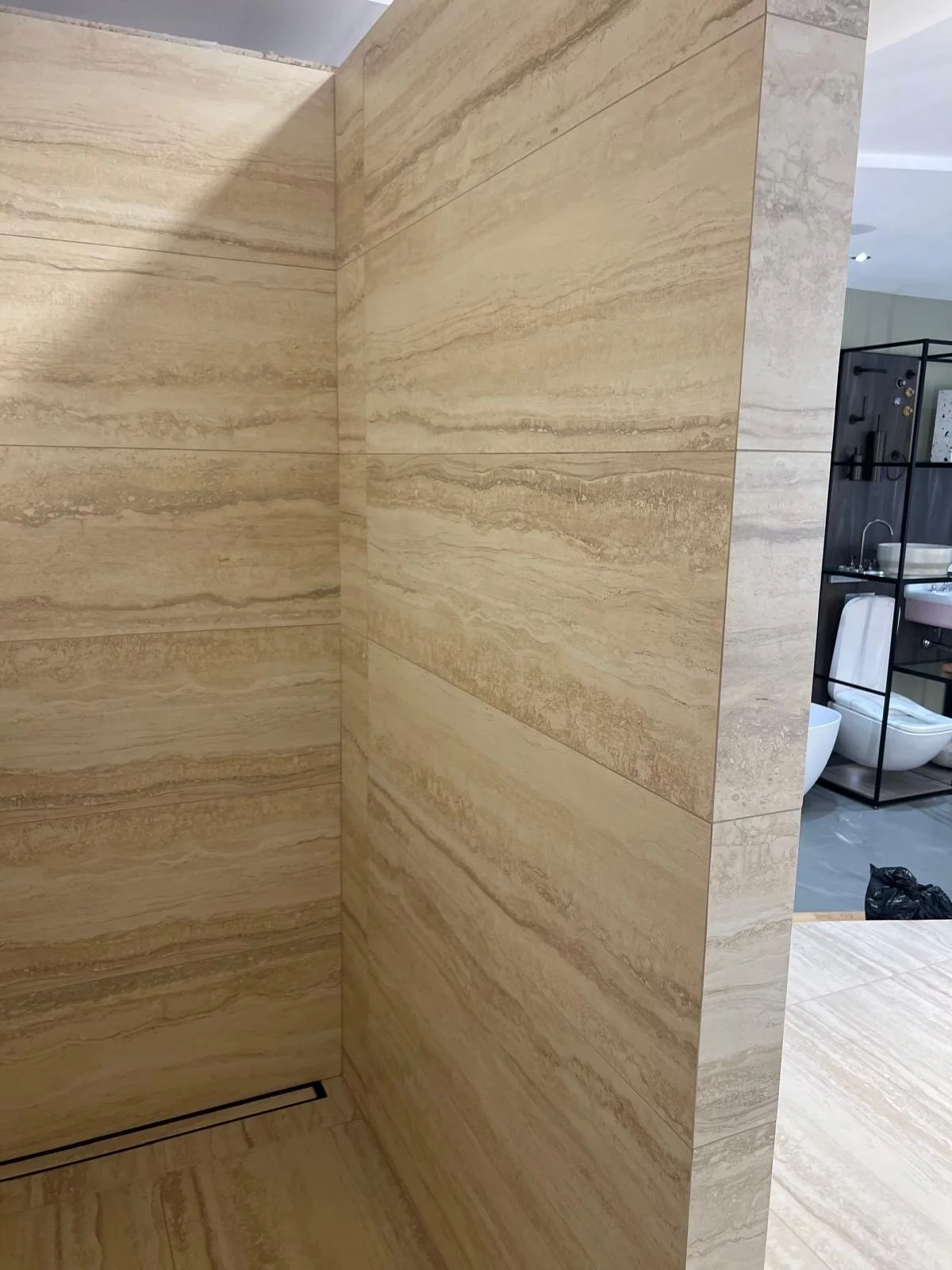 Interior bathroom with beige marble walls and a view of a modern black shelving unit with bathroom fixtures in the background.