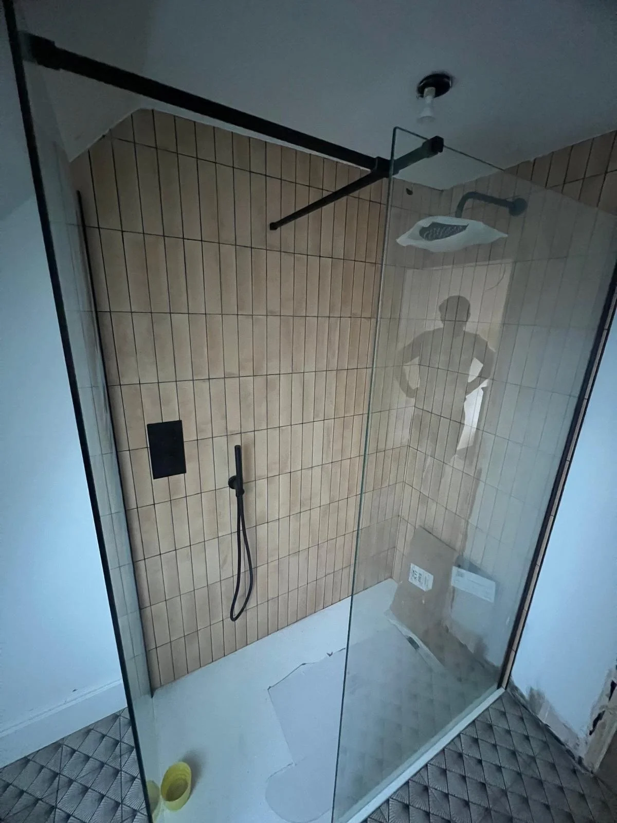 A modern walk-in shower with beige tiles, a black shower head, handheld shower, and a glass partition. Reflects a person taking the photo, with a cracked white floor and a cardboard box outside.