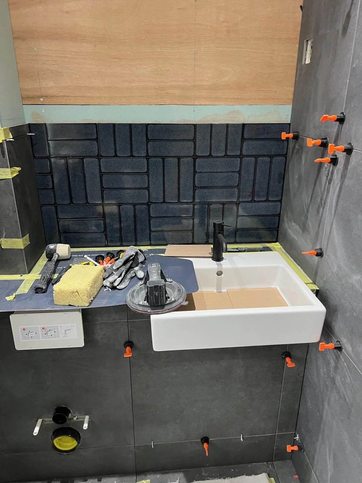 A bathroom under construction with black tile walls, a large white sink, and construction tools and materials like a hammer, sponge, gloves, and spacers. The walls are partially tiled, and some areas are covered with masking tape for installation.